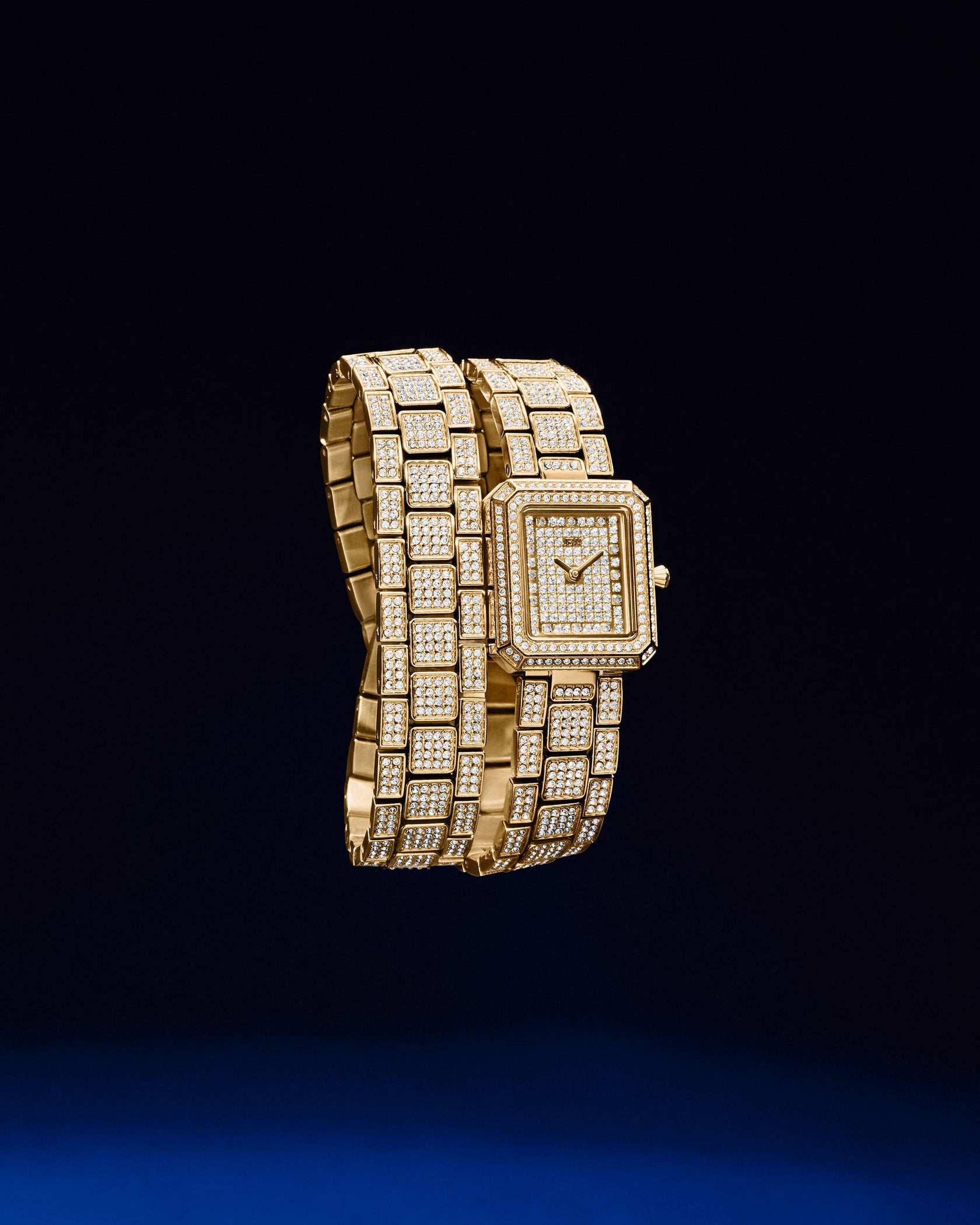 The JBW Watches Arc Double | J6390C features a gold rectangular face and double-wrap diamond-encrusted bracelet with a crystal-studded bezel, shimmering elegantly against a deep blue background.