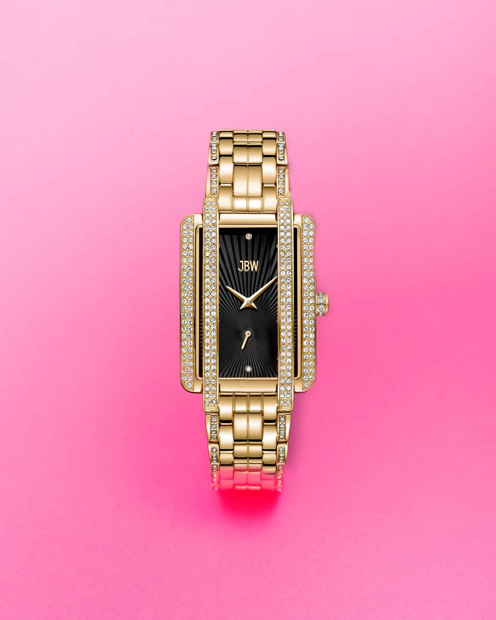 The JBW Mink | J6358G, a gold-plated stainless steel wristwatch with a rectangular case, black face, and diamond accents, is displayed against a vibrant pink background with the JBW logo on the watch face.