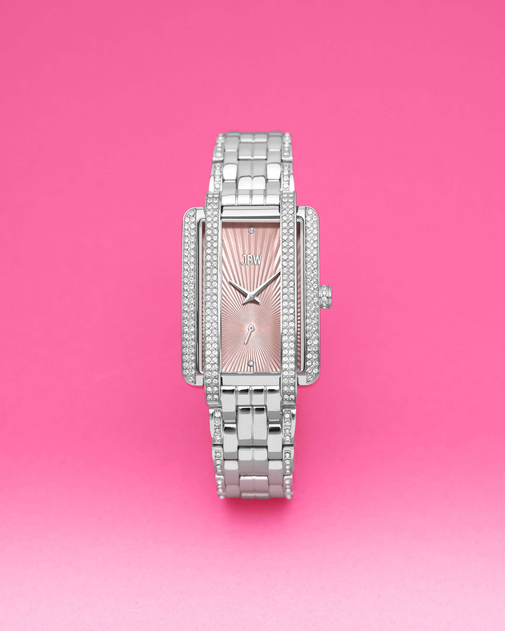 The JBW Mink | J6358H is a silver rectangular case watch with a pink face and diamond accents, standing upright on a bright pink background and paired with a sleek stainless steel bracelet.