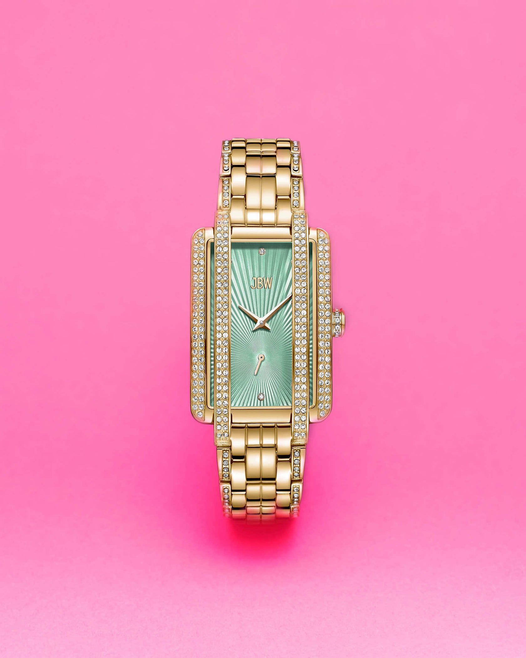 The JBW Mink | J6358J, a gold-plated stainless steel watch with a rectangular green face and diamond accents, stands upright against a bright pink background.