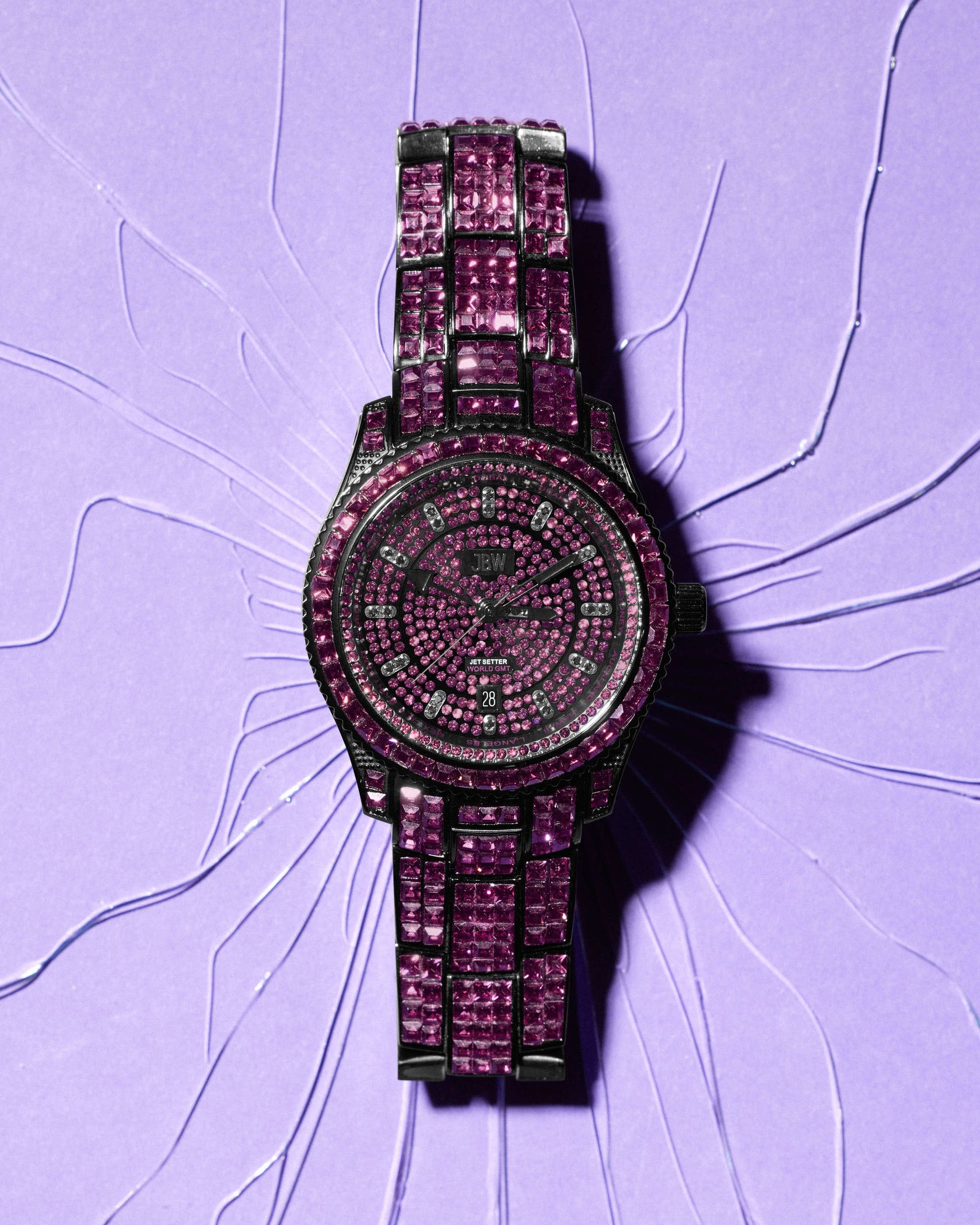 The RESERVE Jet Setter GMT Amethyst | R6393D by JBW Watches dazzles with a watch face and band fully encrusted in brilliant pink stones, showcased against a cracked pastel purple backdrop for bold contrast.