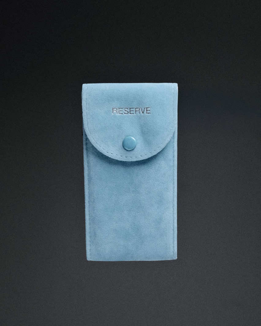A light blue suede pouch with a flap and matching button, embossed with RESERVE, holds your JBW Watches RESERVE Jet Setter GMT Aqua | R6370F—photographed against a dark background.