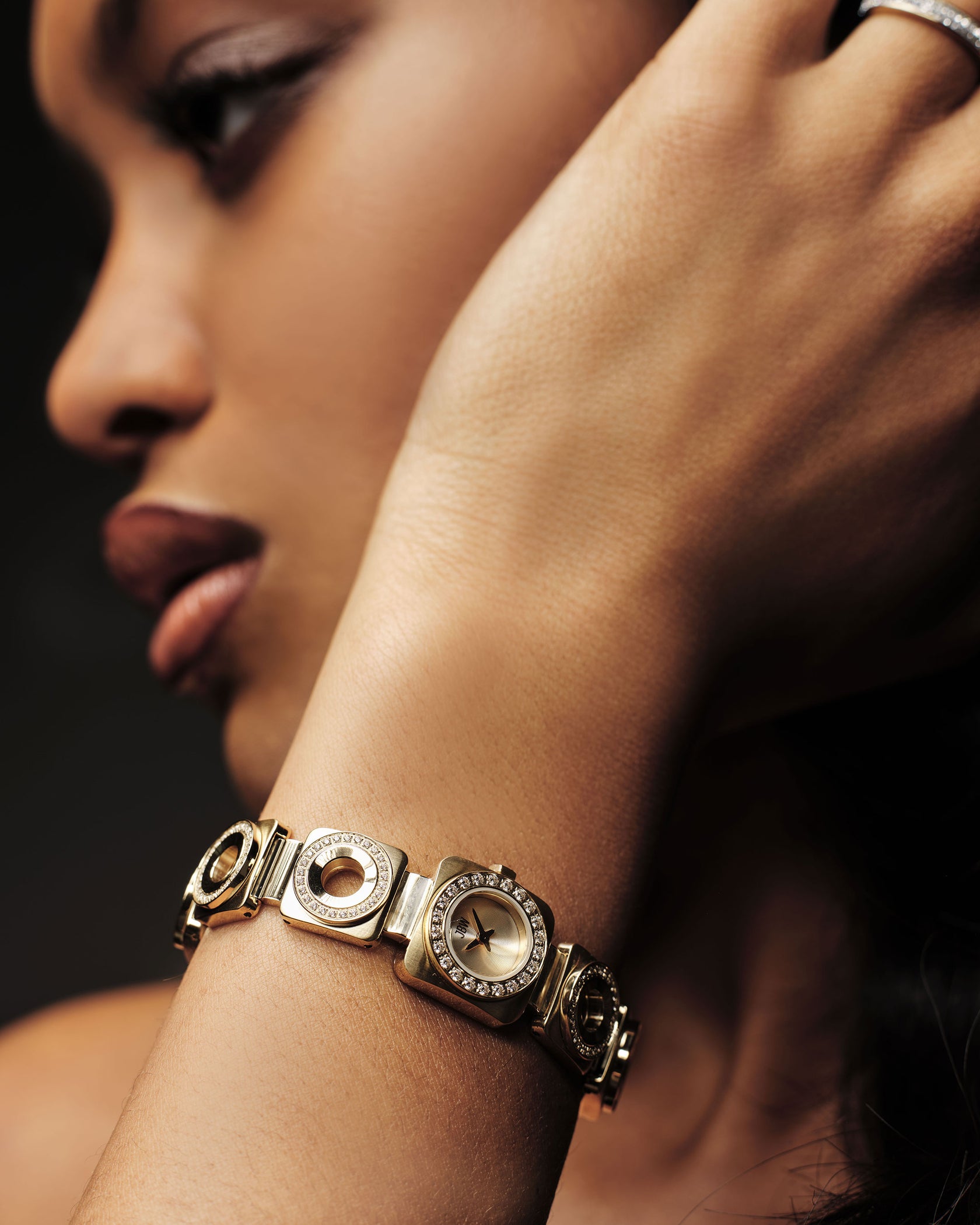 A close-up of a woman’s face and hand highlights the Chasm Petite | J6401A by JBW Watches, an 18k gold-plated watch featuring premium crystals, sparkling details, and Japanese VX movement, complemented by her elegant pose.