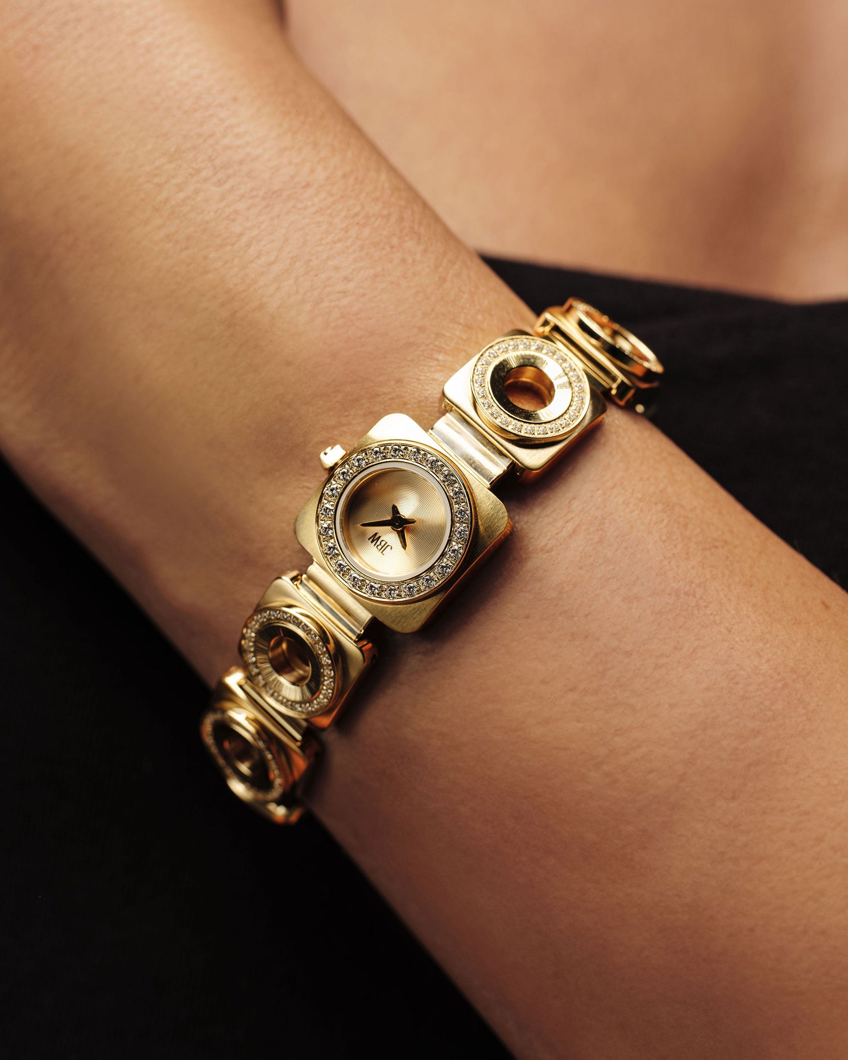 A close-up of the JBW Watches Chasm | J6399A-GA Sample Sale: an 18k gold-plated wristwatch with a circular face, sparkling premium crystals around the dial, and a unique decorative gold band, worn on a wrist against a black background.
