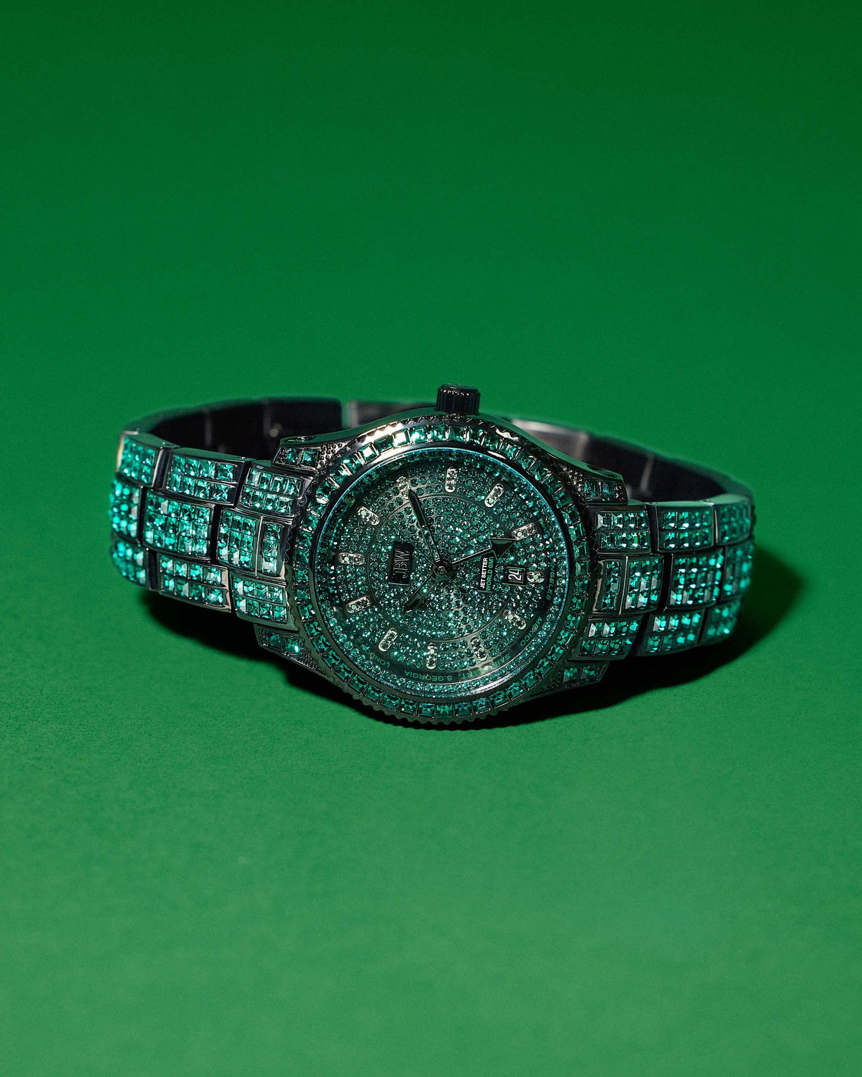 The JBW Watches RESERVE Jet Setter GMT Emerald | R6393C dazzles with green gemstones on the face, bezel, and band, displayed on a solid green background. This luxurious watch features precise Swiss GMT movement.