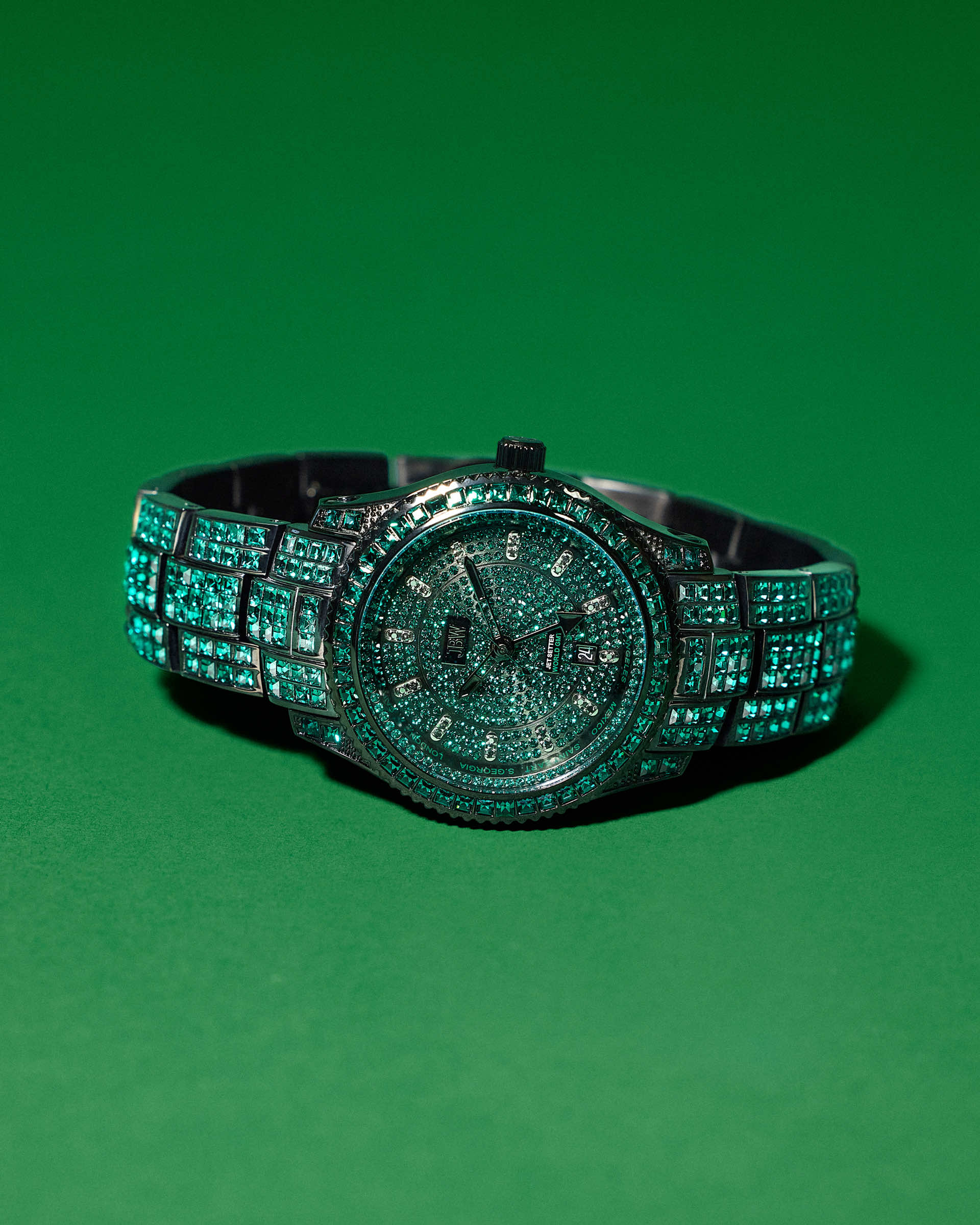 The JBW Watches RESERVE Jet Setter GMT Emerald | R6393C dazzles with green gemstones on the face, bezel, and band, displayed on a solid green background. This luxurious watch features precise Swiss GMT movement.