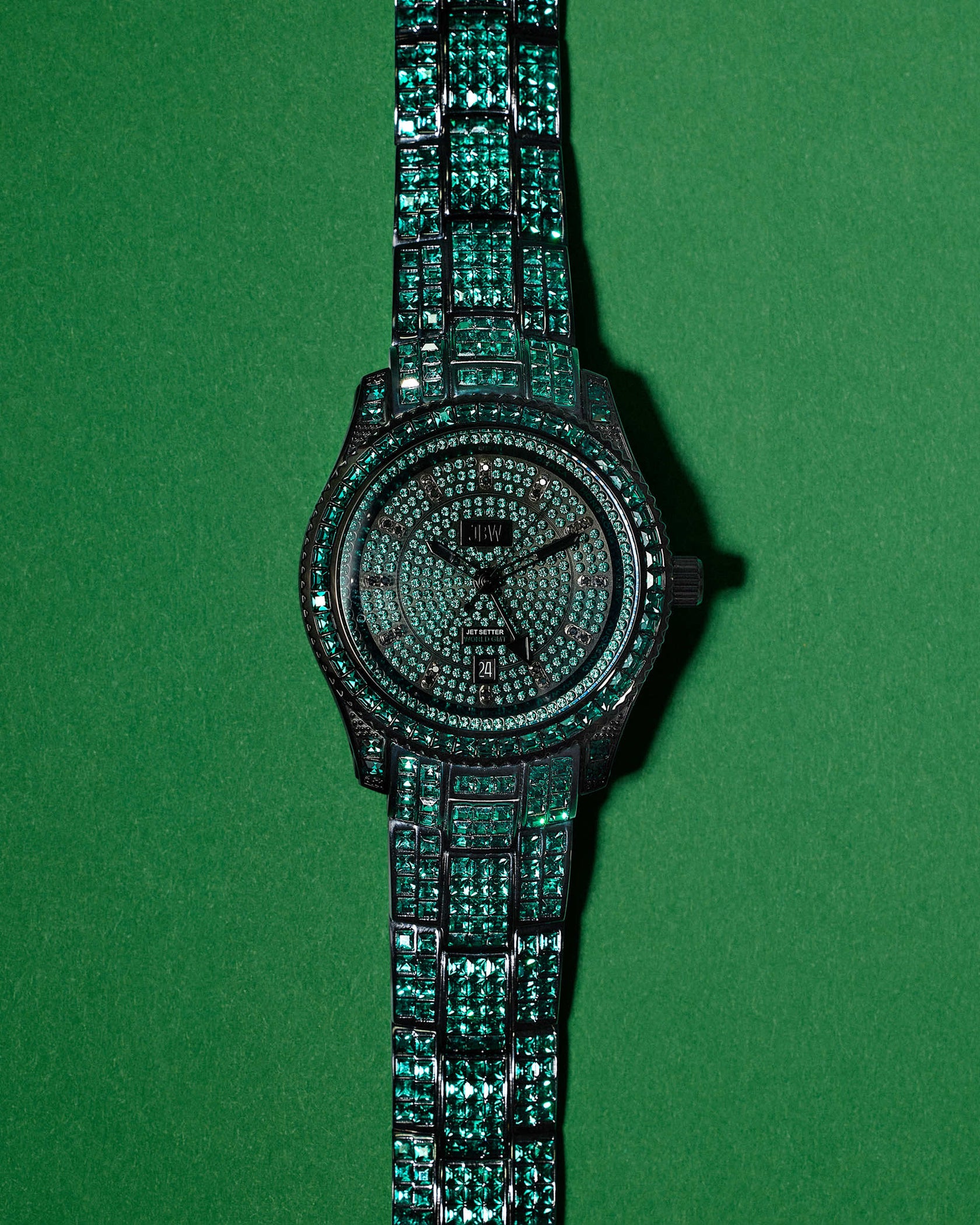 The JBW Watches RESERVE Jet Setter GMT Emerald (R6393C) dazzles on a green backdrop, highlighting its diamond and emerald-encrusted face and band, along with precise Swiss GMT movement and intricate detailing.