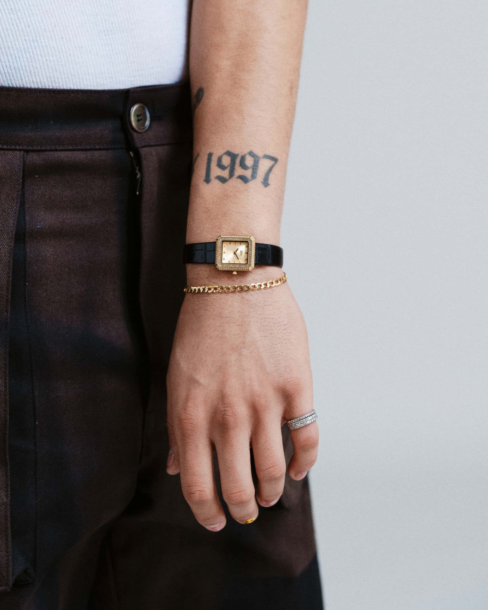 A person’s left hand and wrist wear the JBW Watches Arc Leather | J6390LA, a gold unisex watch with a black leather band, paired with a gold chain bracelet, silver ring, and visible 1997 tattoo. The person is also wearing dark pants.