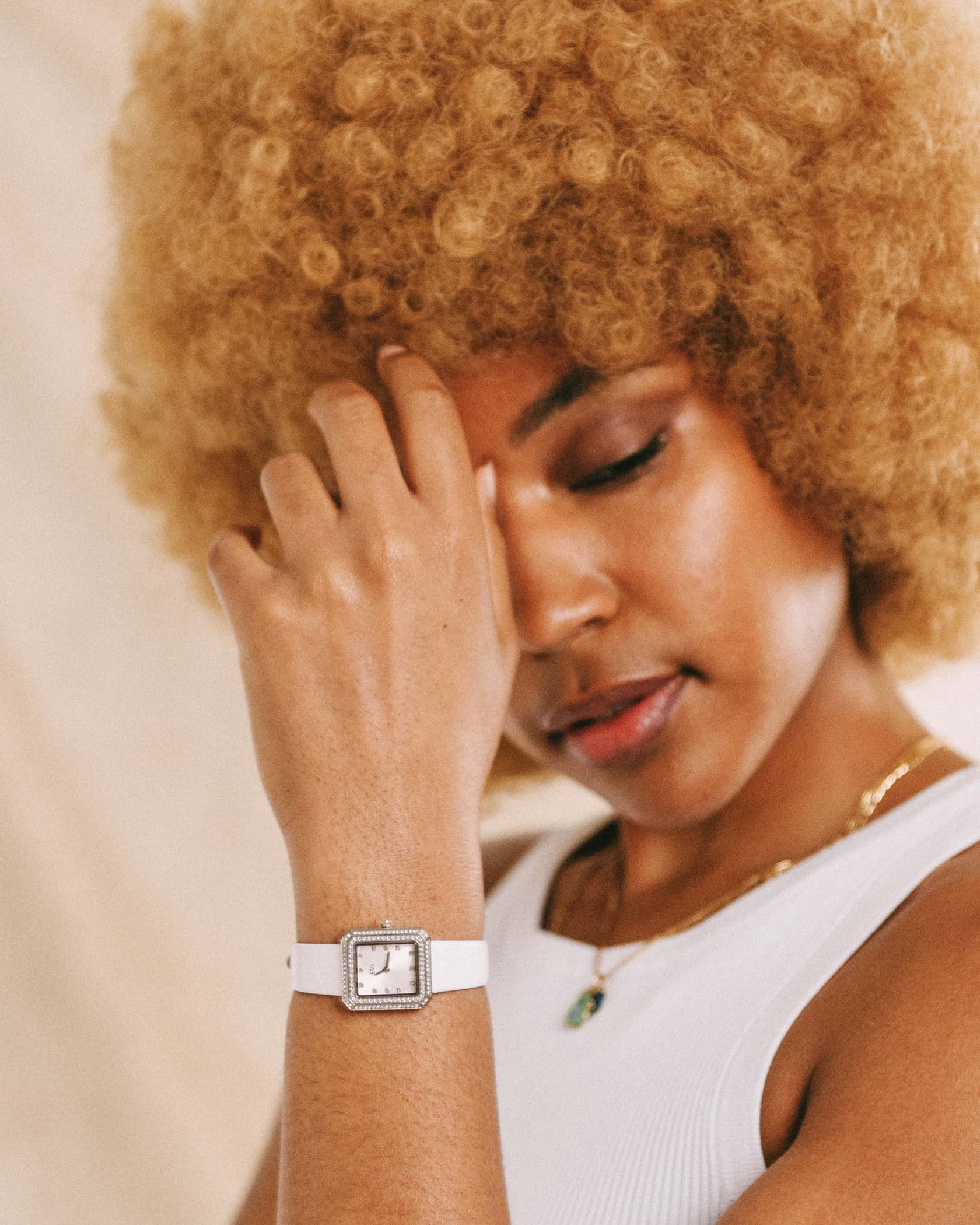 A woman with curly blonde hair in a white tank top touches her forehead gently, eyes closed in serenity, wearing the JBW Watches Arc Leather | J6390LC square-faced unisex watch with a leather band.