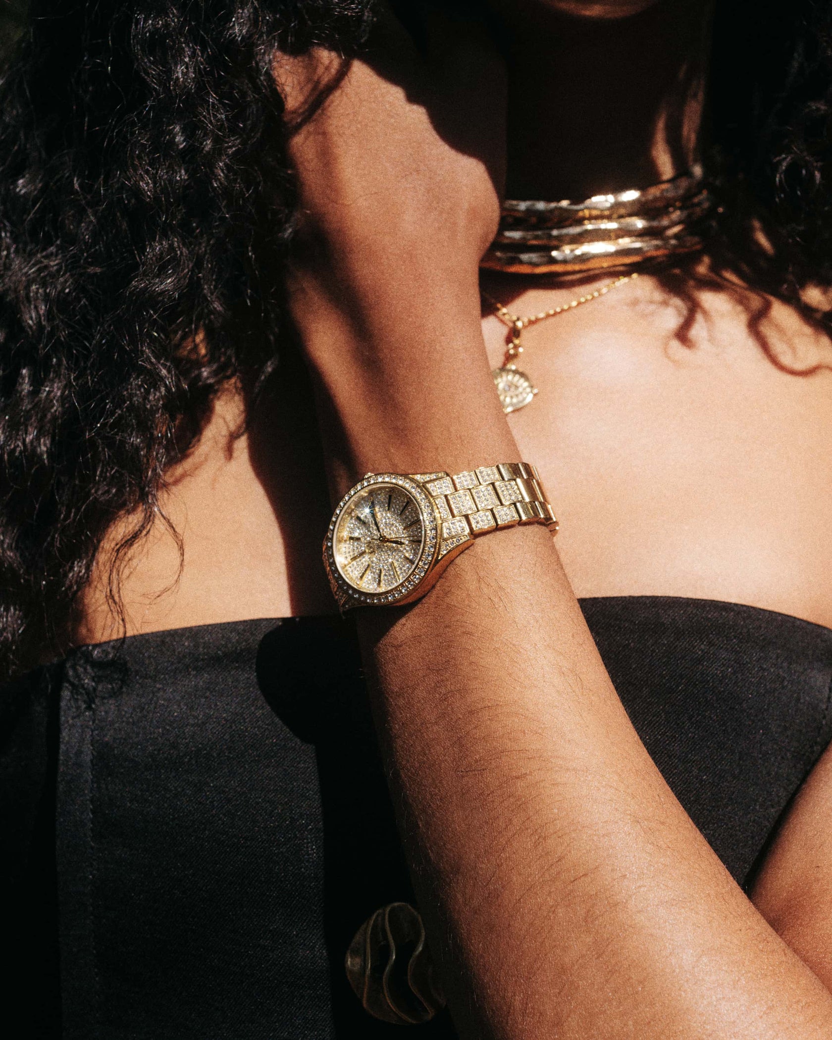 Wearing the JBW Cristal 34 | J6383A diamond watch and layered gold necklaces, someone with curly hair and a black strapless top shines in sunlight that highlights their jewelry and skin.