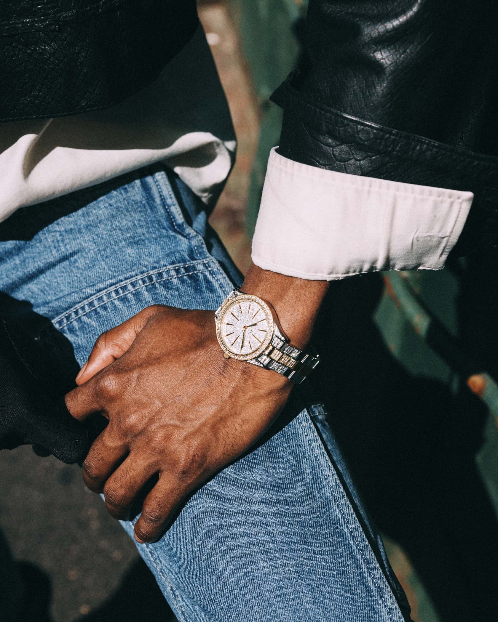 A close-up of a person's hand and wrist wearing the JBW Cristal 34 | J6383D diamond watch, resting on their knee. They're dressed in blue jeans, a white shirt, and a black leather jacket.