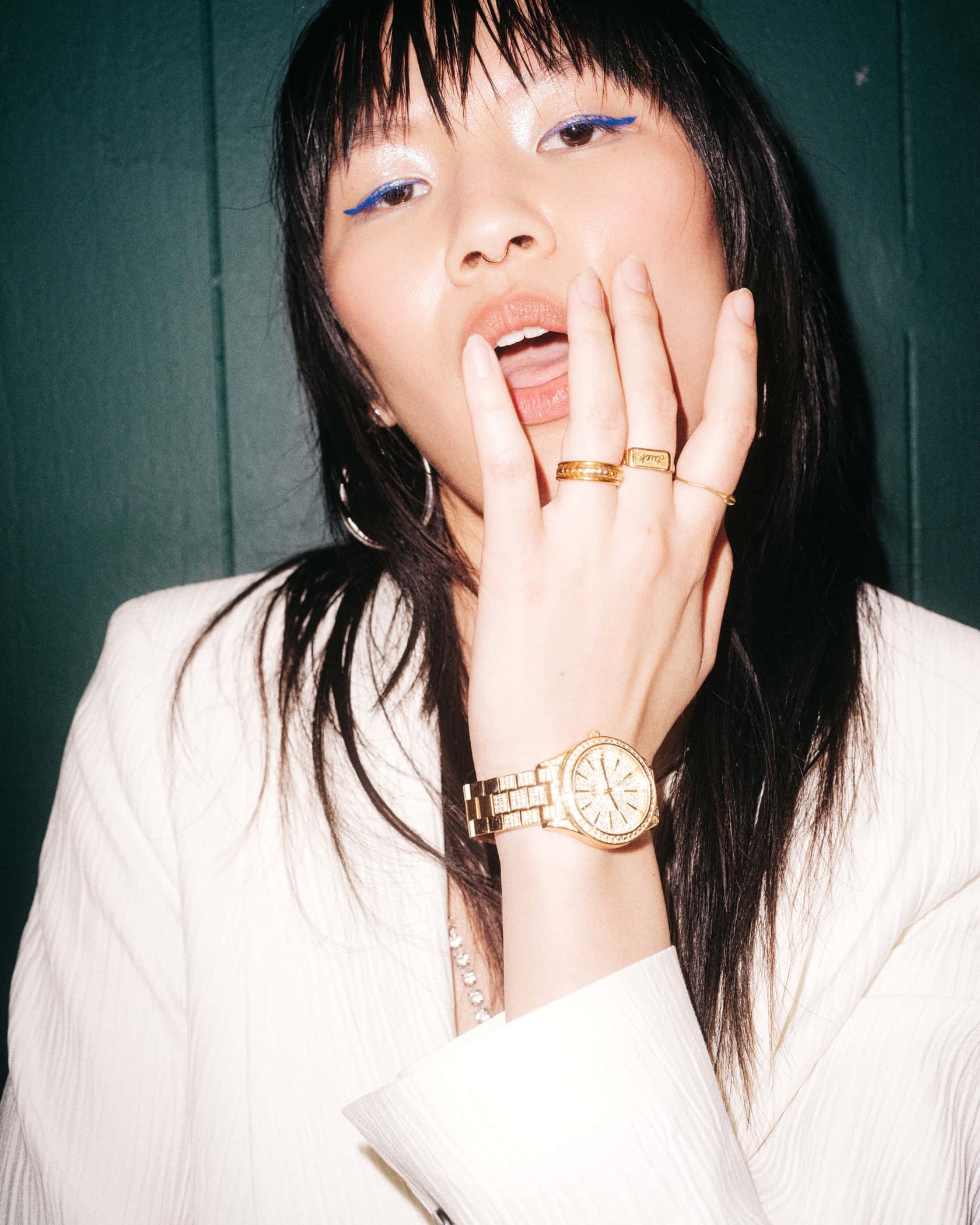 A woman with long dark hair, blue eyeliner, and white attire poses against a green background, wearing the JBW Cristal 39 | J6346A gold-plated stainless steel watch and rings as she touches her lips.