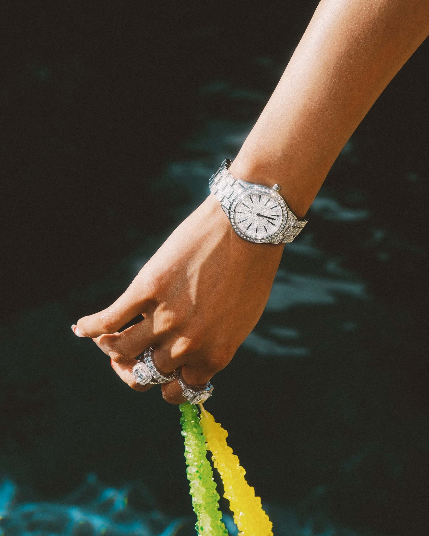 A close-up of a hand wearing the JBW Cristal 39 (Sample Sale) | J6346C-GA (Grade A) stainless steel, diamond-encrusted watch holding green and yellow rock candy sticks against a dark, blurred background.