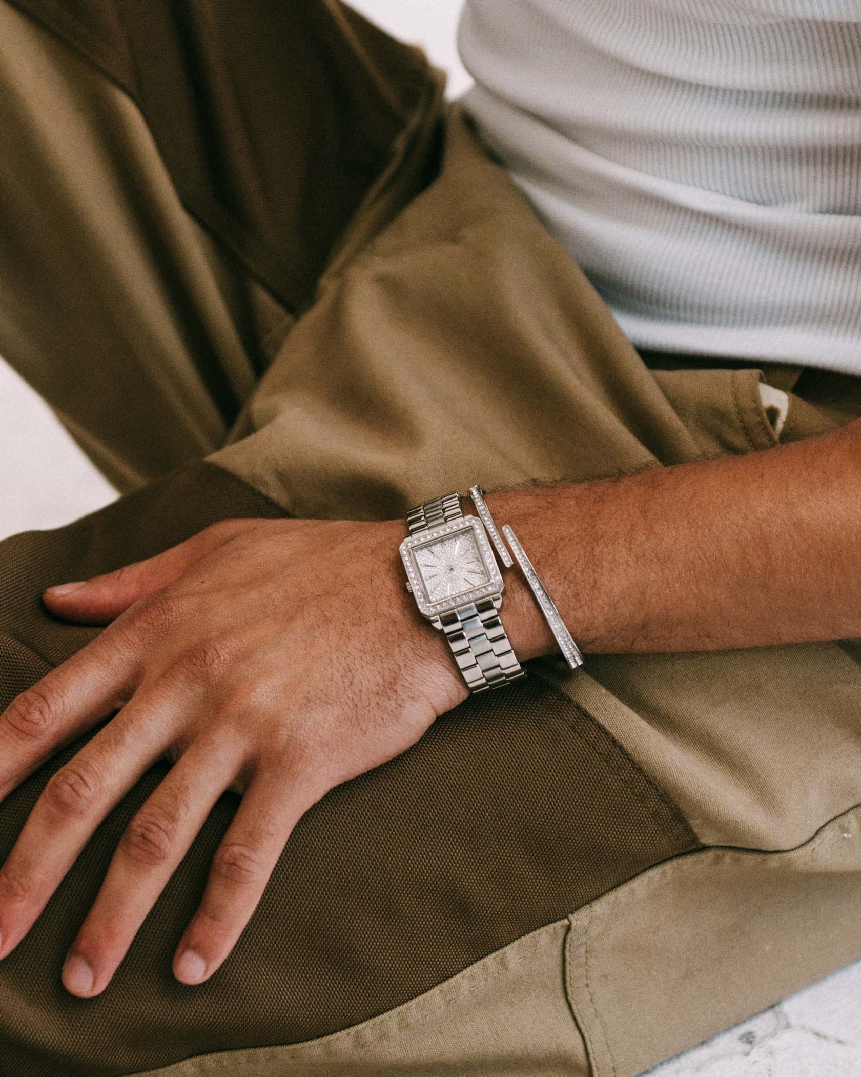 A person wearing brown pants and a white ribbed tank top rests their left hand on their knee, showcasing the JBW Cristal 28 | J6387-SetC—a square-faced silver watch adorned with Austrian crystals and a slim silver bracelet.