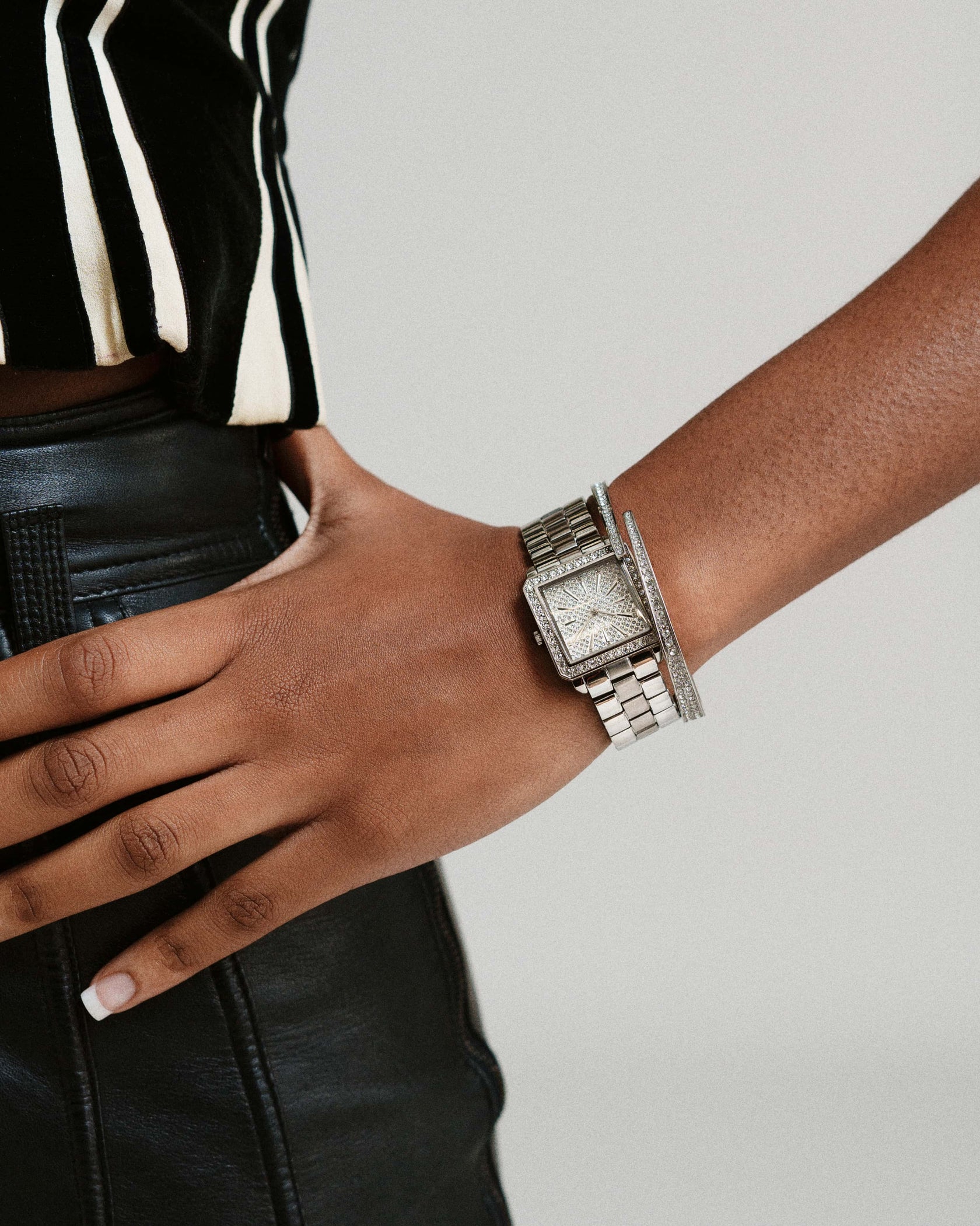 A person wearing black leather pants, a black-and-white striped top, and the JBW Cristal 28 | J6387-SetC diamond watch with a matching silver bracelet rests their hand on their hip against a light background.