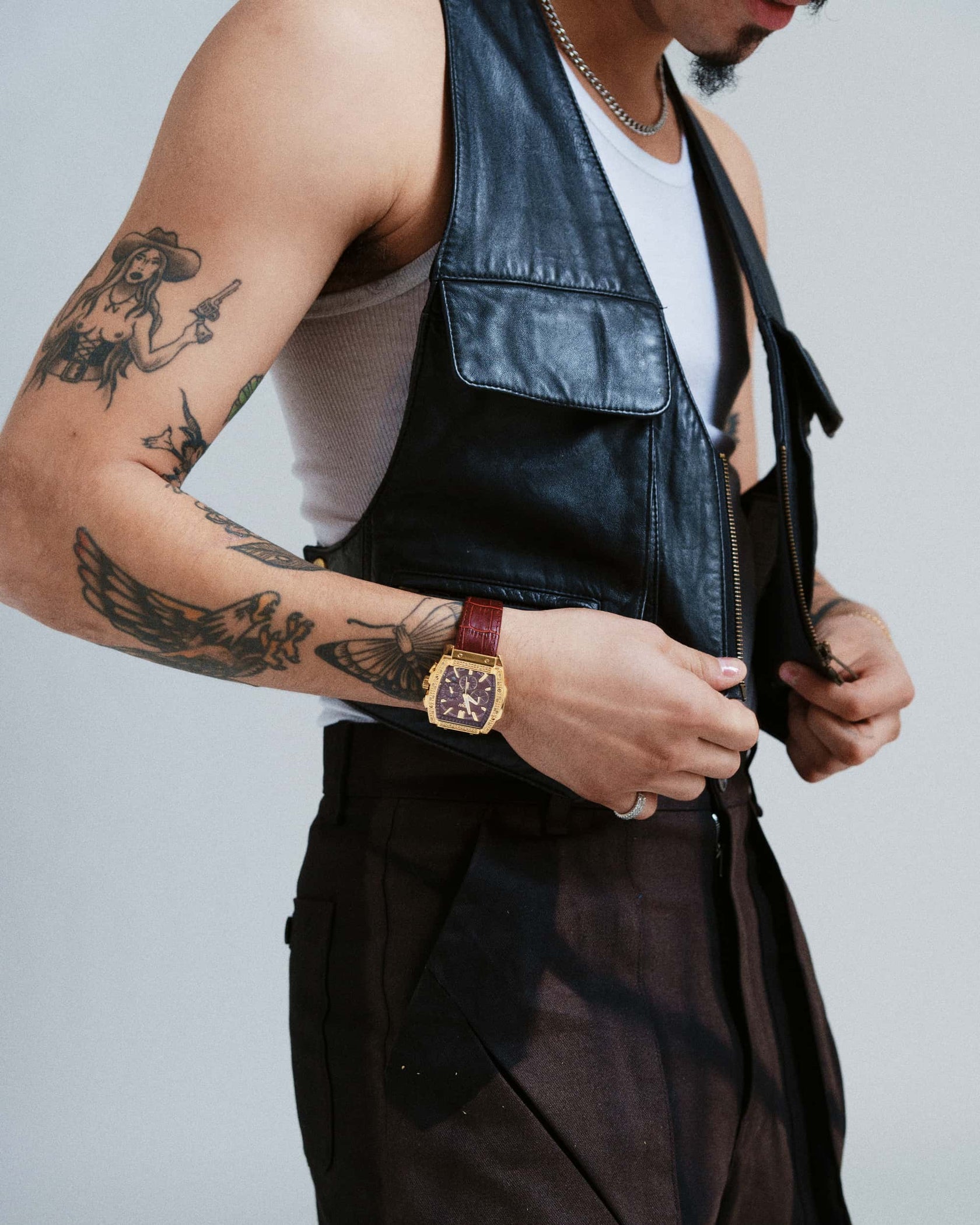 A person in a sleeveless leather vest over a white tank top and dark pants adjusts the vest’s zipper, revealing tattoos and wearing a JBW Echelon | J6379B diamond chronograph watch with a brown band plus a chain necklace.