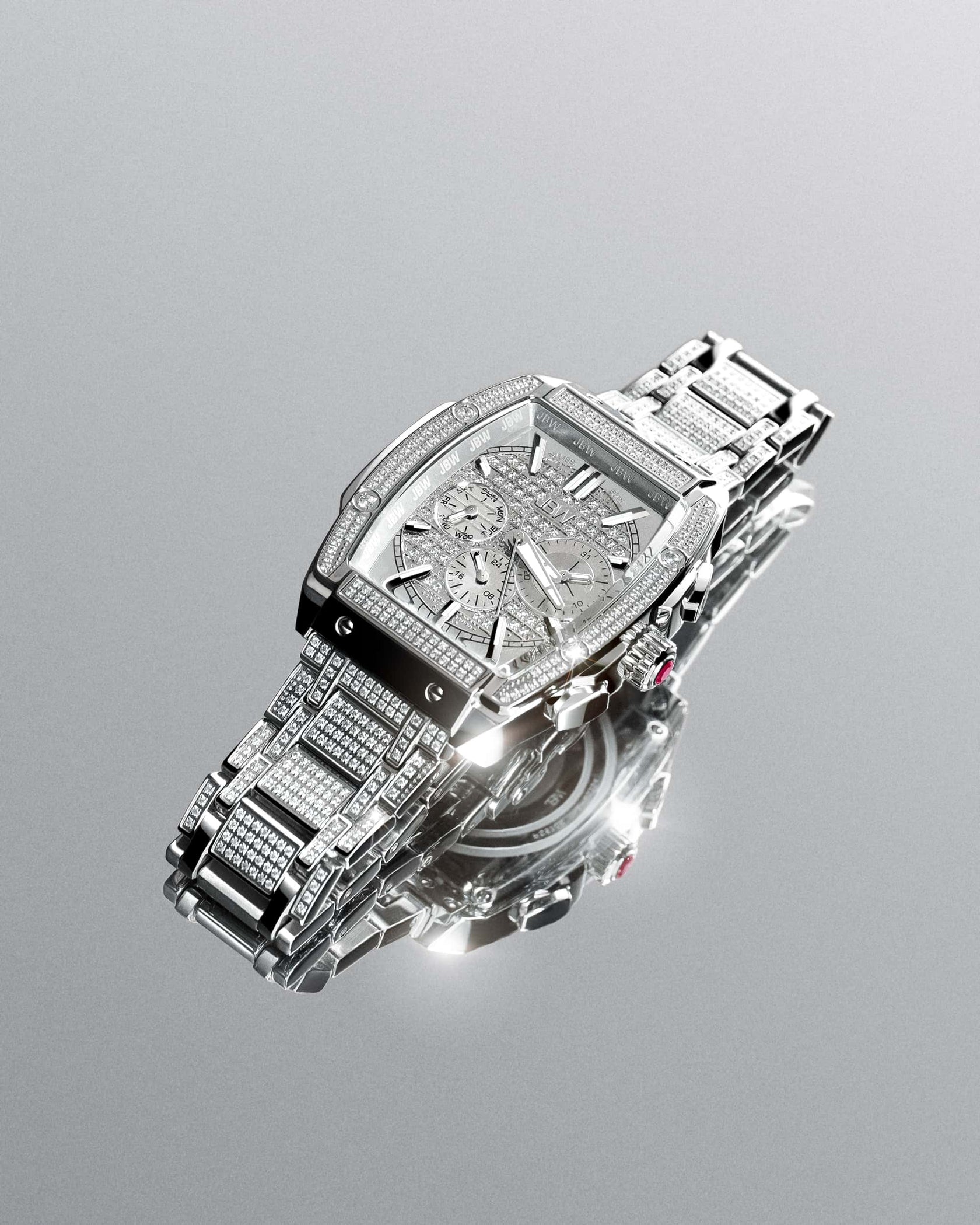 The JBW Echelon PS | PS570B, a luxurious 18K gold-plated watch with diamond accents, Roman numerals, and a silver metal band, is displayed elegantly on a reflective surface.