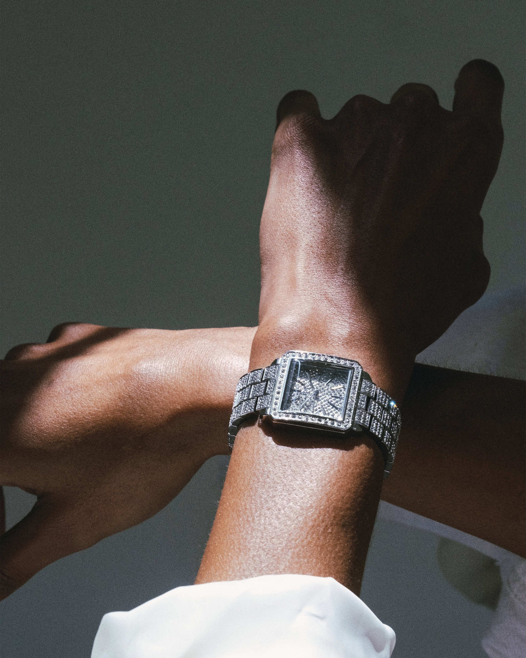 A close-up shows a wrist wearing the JBW Cristal 28 | J6386C-GA Sample Sale (Grade A) diamond watch with a square face and Swiss movement. The other hand crosses above it, set against a dark, shadowy background.