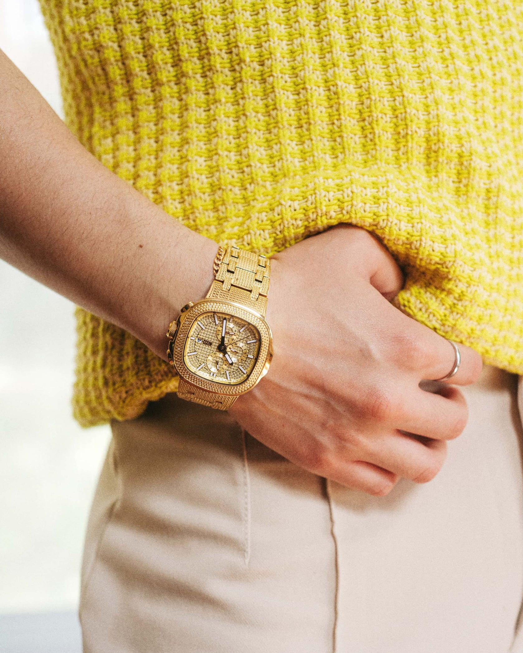 Wearing a yellow textured sweater and light pants, the person rests their right hand on their hip, showing off the JBW Heist | J6380A—an 18K gold-plated stainless steel wristwatch embellished with sparkling stones.