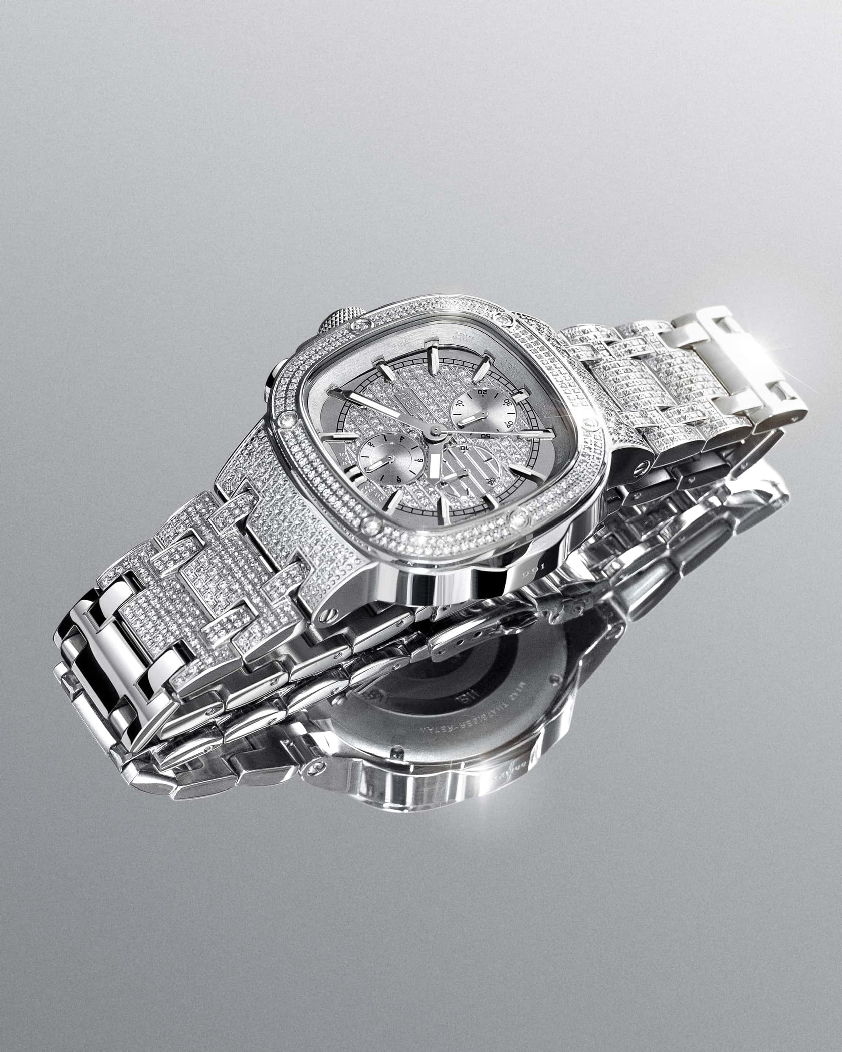 The JBW Watches Heist PS | PS545B, a luxury Swiss chronograph with a diamond bezel, is showcased on a reflective surface, highlighting its intricate stainless steel bracelet, detailed dial, and sparkling accents.