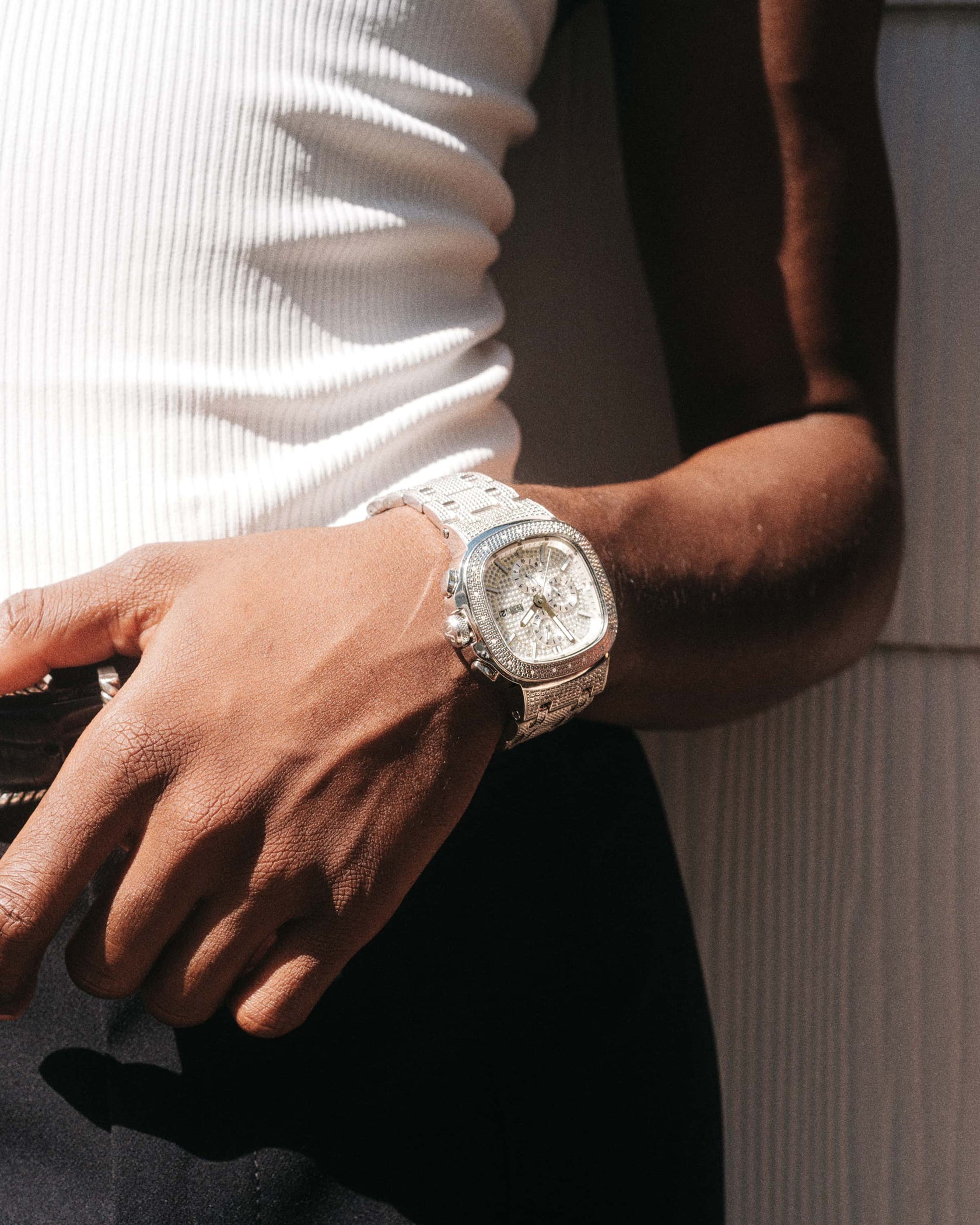 A person in a white ribbed tank and dark pants stands with one hand on their waistband, showcasing the JBW Heist | J6380D diamond watch with a stainless steel bracelet on their left wrist.