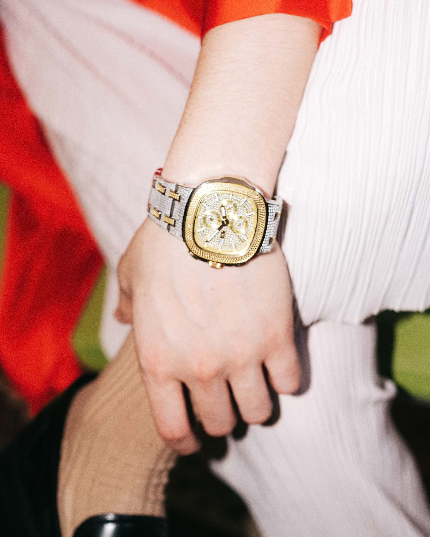 A close-up of a wrist adorned with the JBW Heist | J6380B womens luxury gold-plated stainless steel watch, featuring a detailed face. The person wears white pants, a red top, and black shoes while resting their arm on a bent knee.