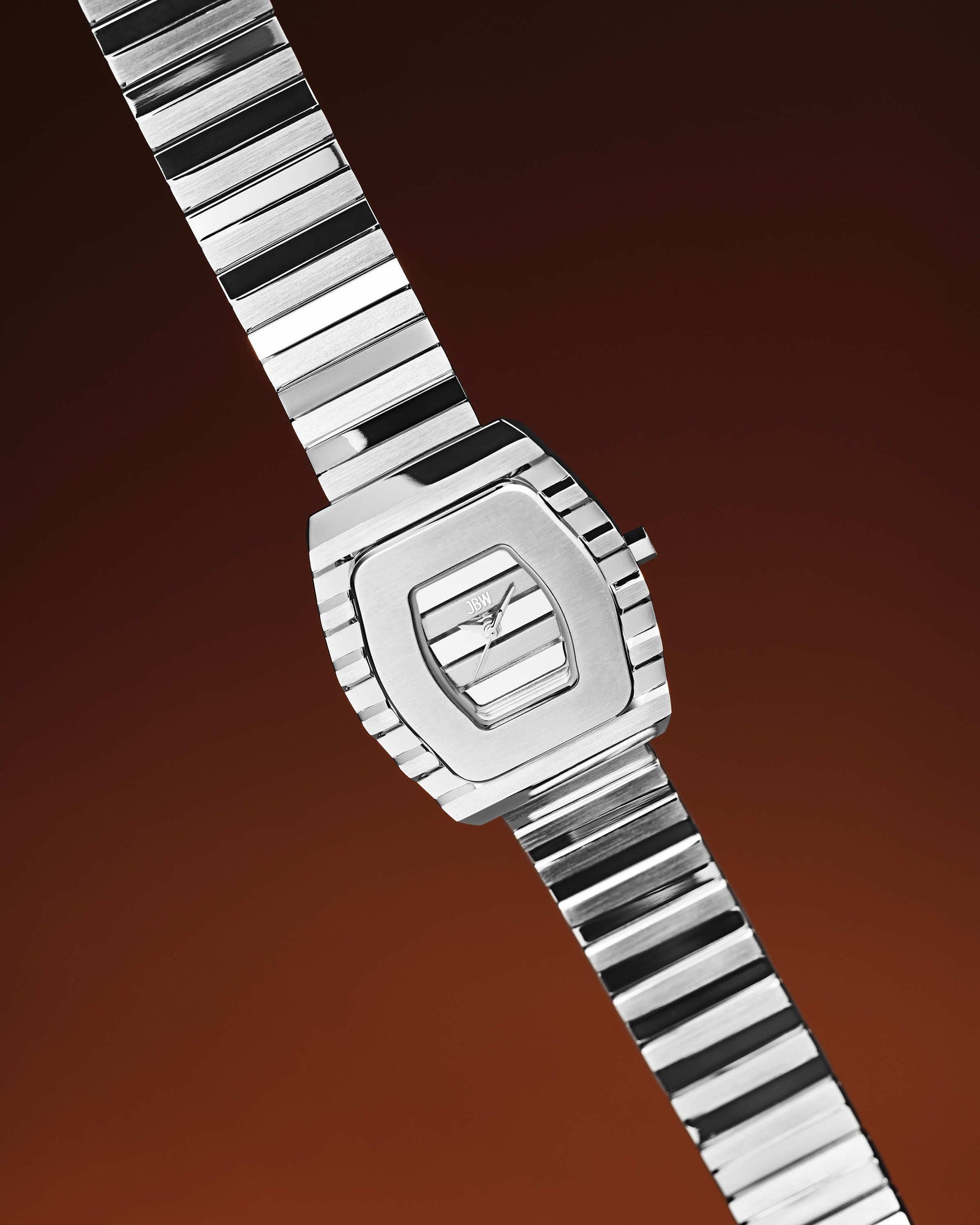 The JBW Watches Atlas | J6403B is a modern women’s stainless steel bracelet watch with a rectangular face and metal link band, featuring Swiss Ronda movement, displayed diagonally on a dark brown gradient background.