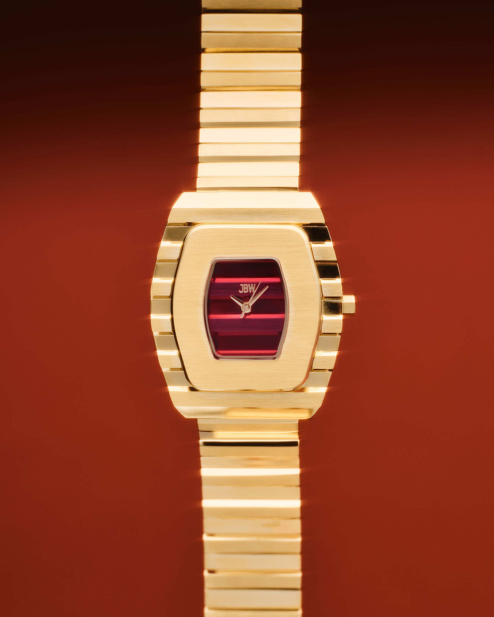 The JBW Watches Atlas | J6403E, an 18K gold-plated womens watch with a rectangular case and red dial, appears centered against a warm reddish-brown background. Its polished metal band and JBW branding add to its luxury appeal.