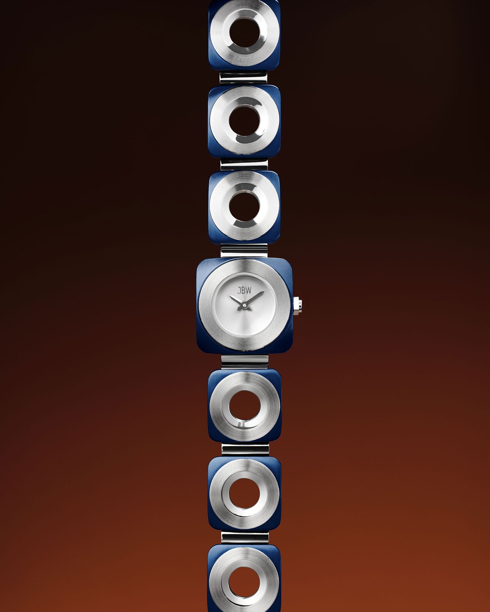 The Chasm | J6398C by JBW Watches is a minimalist wristwatch with a silver circular face and blue-silver square links, finished with two-tone IP blue plating. Displayed vertically on a gradient brown background, it has no numerals or markings.