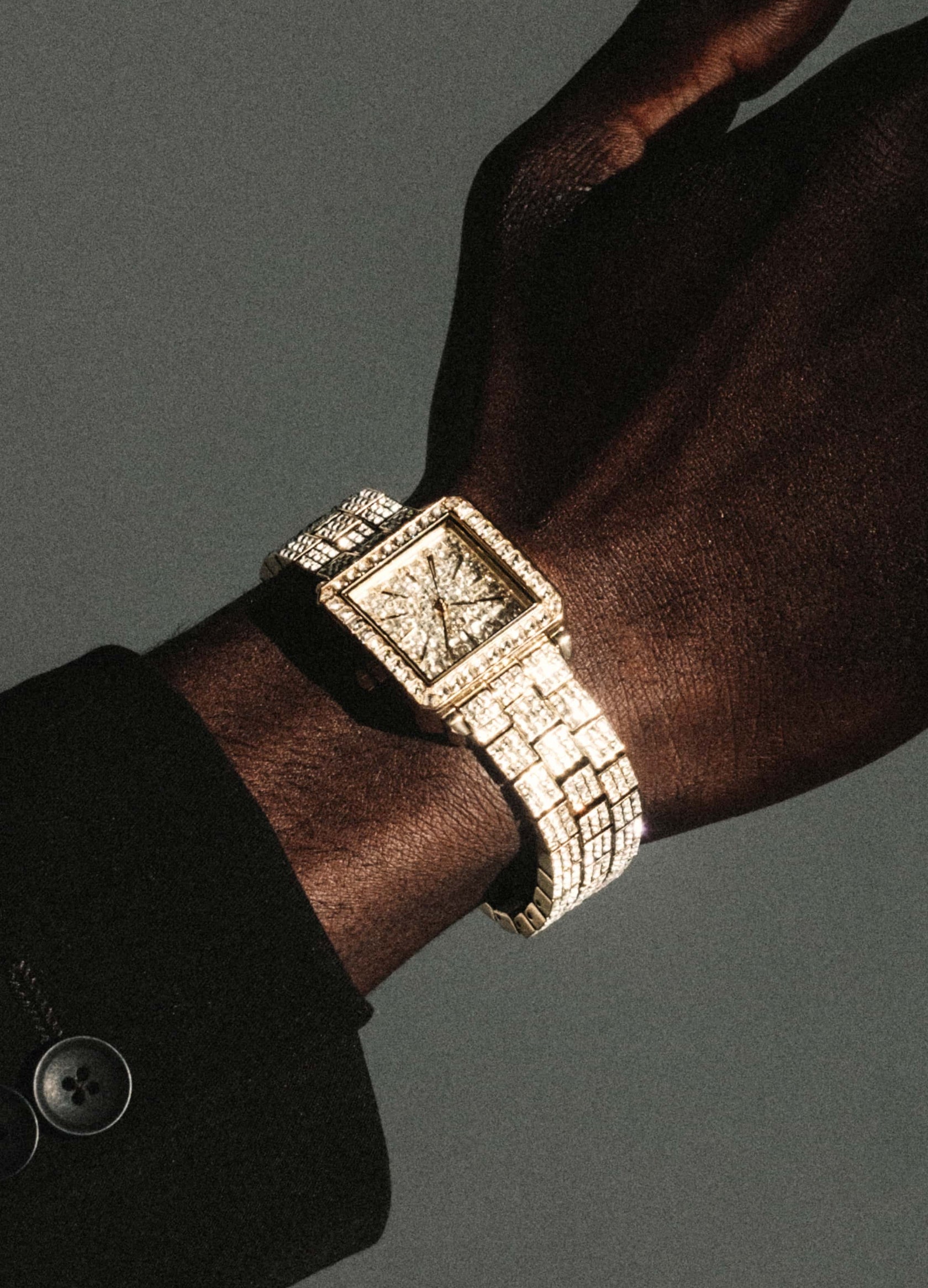 A close-up shows a wrist wearing the JBW Watches RESERVE Cristal Gold | R6386A, featuring diamond hour markers on a gold, diamond-encrusted square face. A dark sleeve and button appear against a neutral gray background.