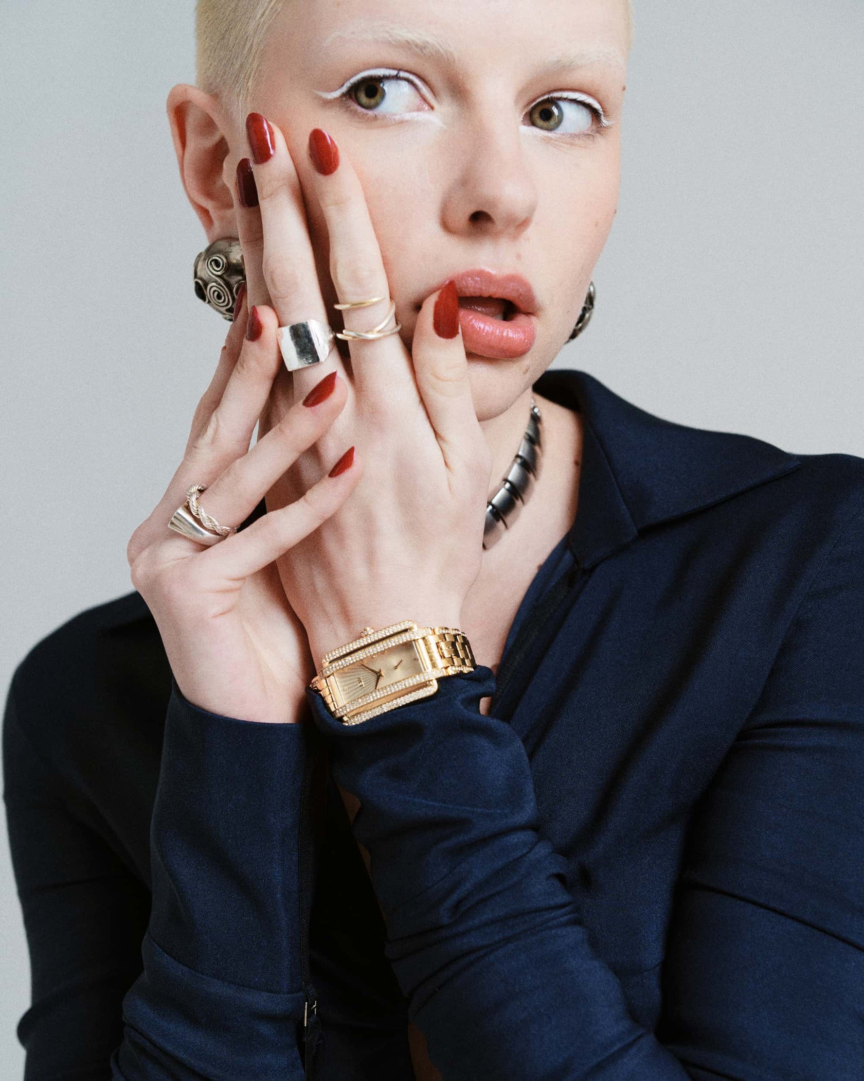 A person with short blond hair and red nails poses with their hands near their face, wearing chunky rings, large earrings, a dark blue top, and the JBW Mink | J6358B gold-plated watch.
