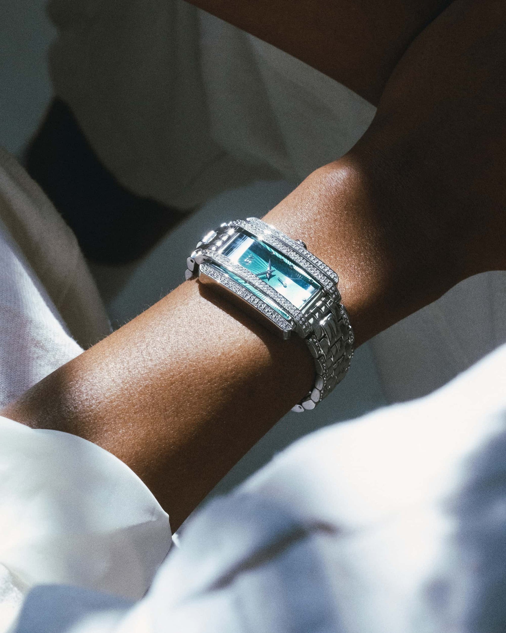 A close-up of a wrist wearing the JBW Mink | J6358A, a sleek silver rectangular watch with a crystal-embellished stainless steel bracelet and blue face, sparkling against soft light and white clothing.