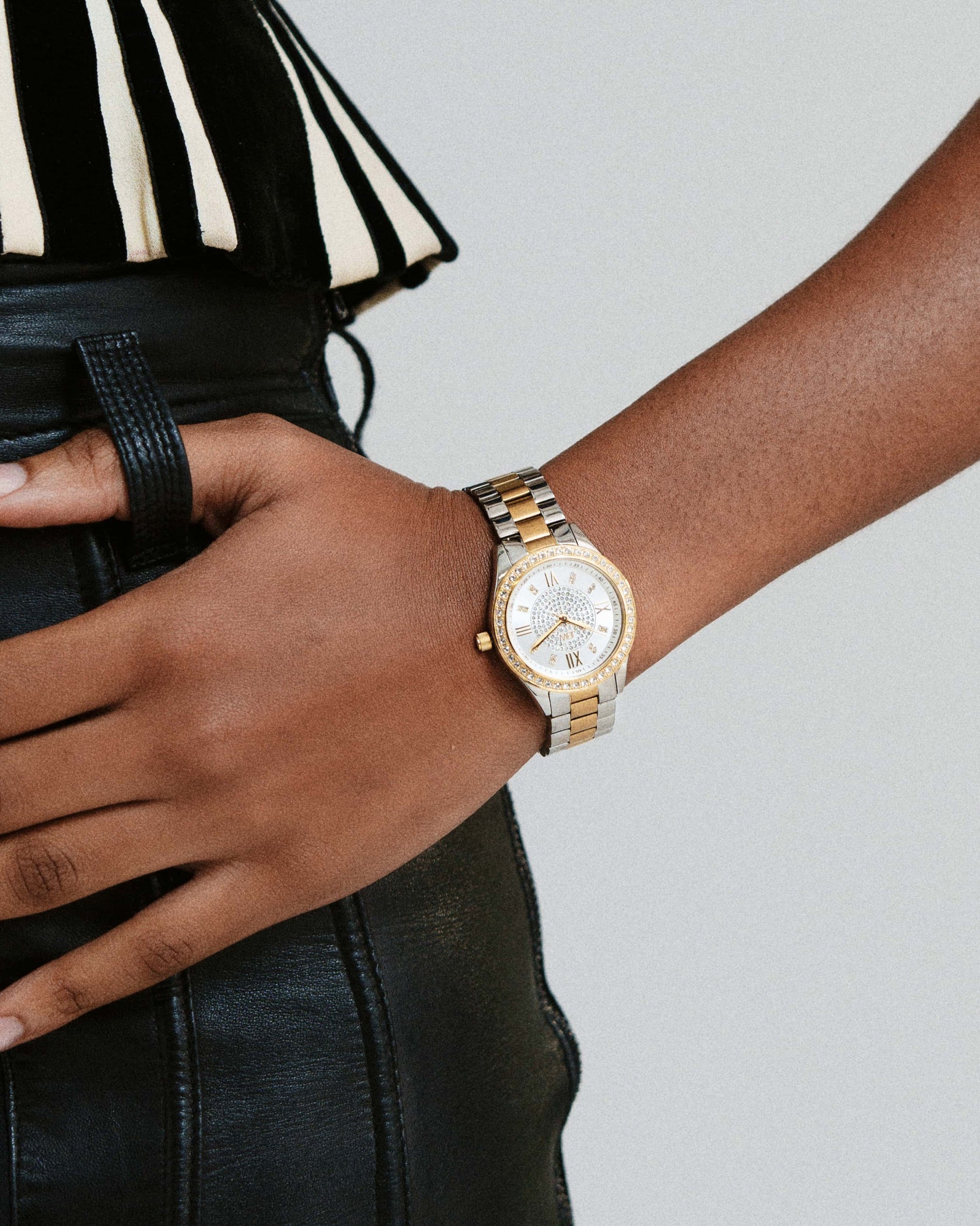 A person in black leather pants and a striped black-and-white top rests their hand on their hip, wearing the JBW Mondrian | J6303G gold-plated stainless steel wristwatch with a white face.