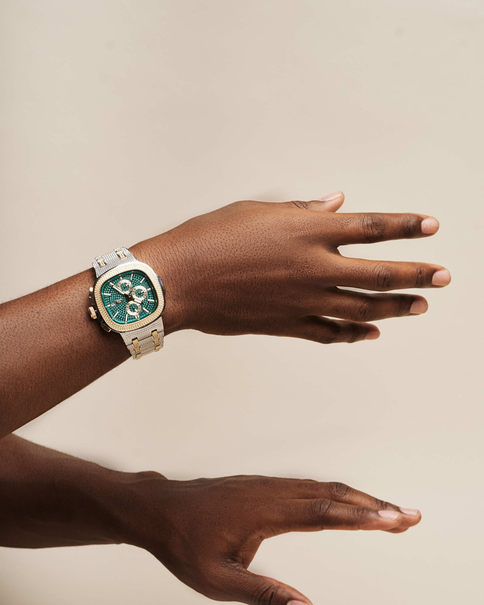 A person with brown skin displays a JBW Watches Heist | J6380F gold-plated stainless steel wristwatch on one hand over the other against a plain beige background.