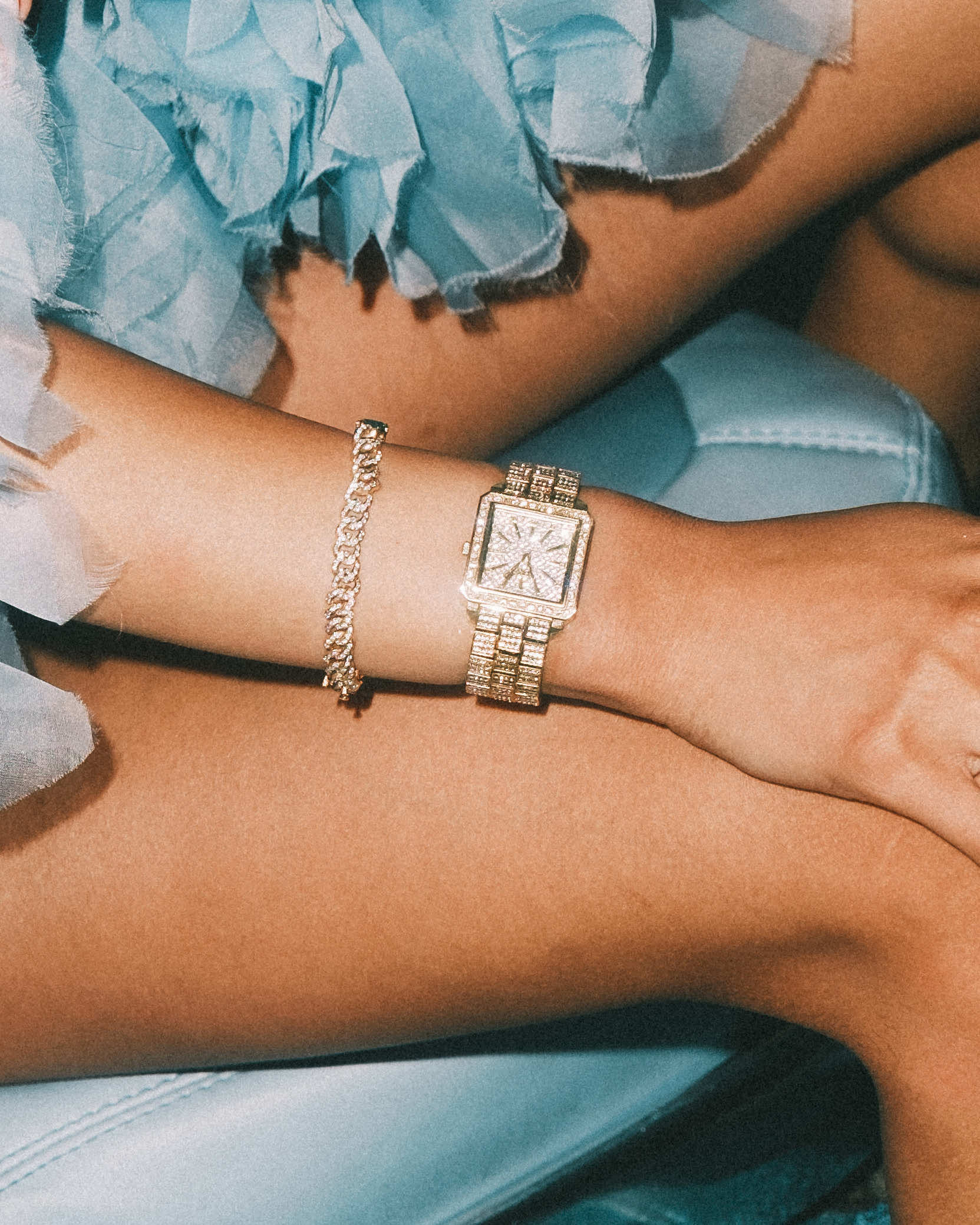 A close-up of crossed arms resting on a light blue surface, showcasing the JBW Cristal 28 | J6386A gold-plated stainless steel wristwatch and a matching bracelet, with a frilly blue garment partially visible.