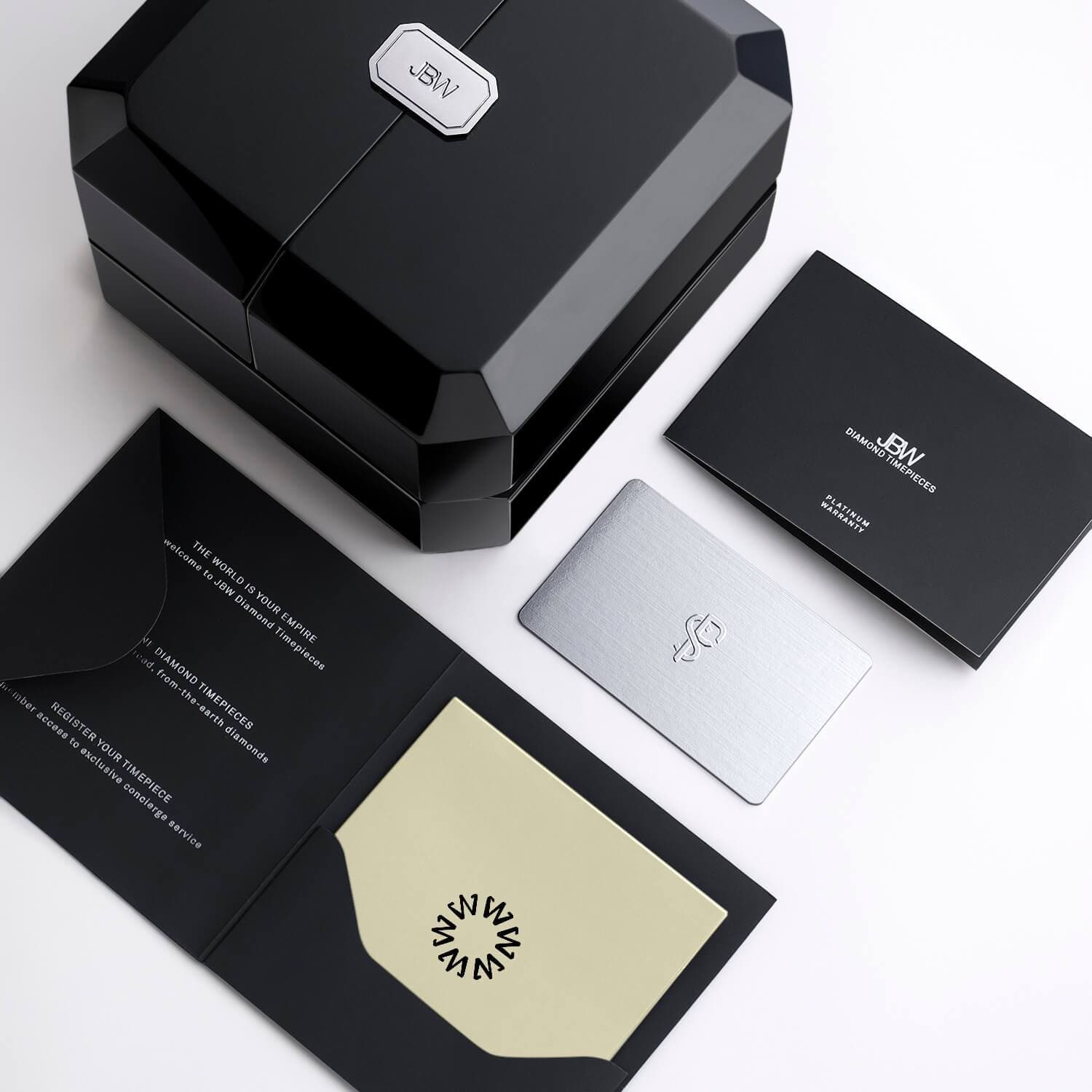 A sleek black box with a silver logo, a matching black envelope, silver card, and gold-accented brochure are elegantly arranged to showcase the JBW Watches Heist PS | PS545A gold-plated stainless steel watch on a pristine white surface.