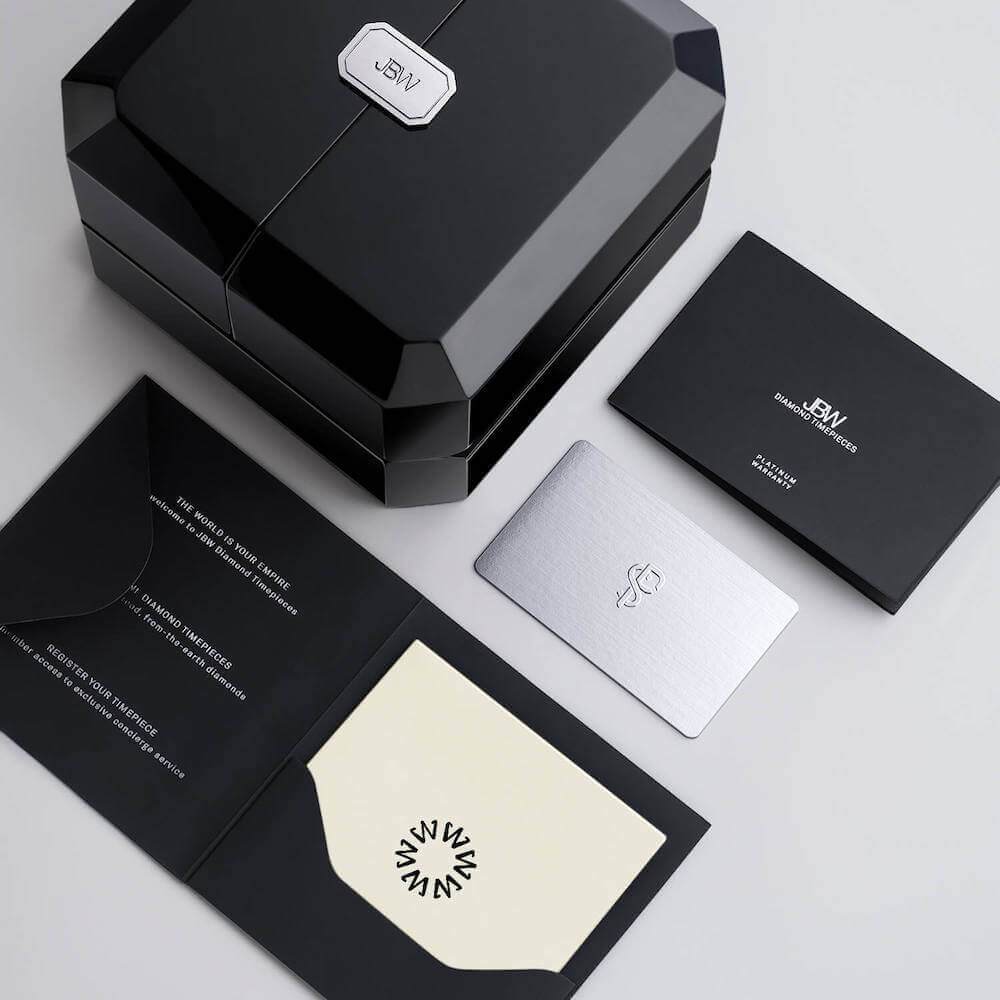 A black, faceted JBW Watches gift box with a metallic logo sits beside a black envelope, silver card, and cream folded card—an elegant display for the Jet Setter PS (PS550B-GB Sample Sale, Grade B) diamond Swiss watch on a light gray surface.