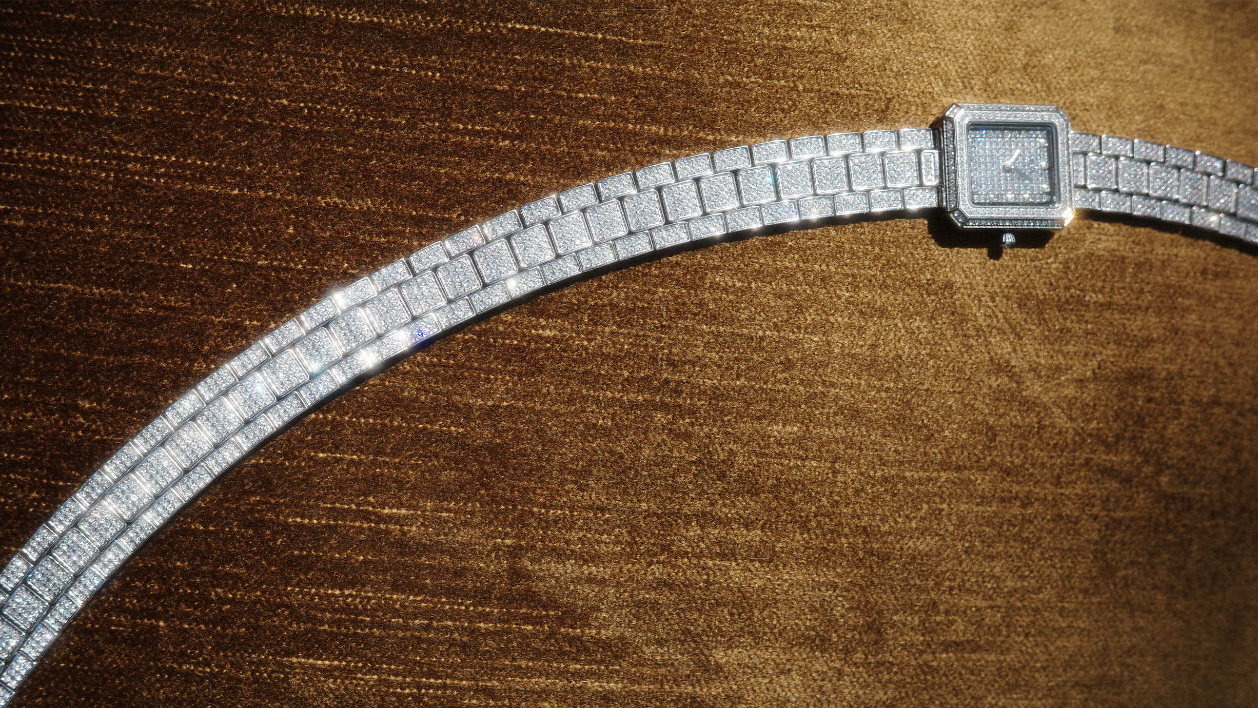 A diamond-encrusted wristwatch with a square face is displayed against a shimmering brown textured background.