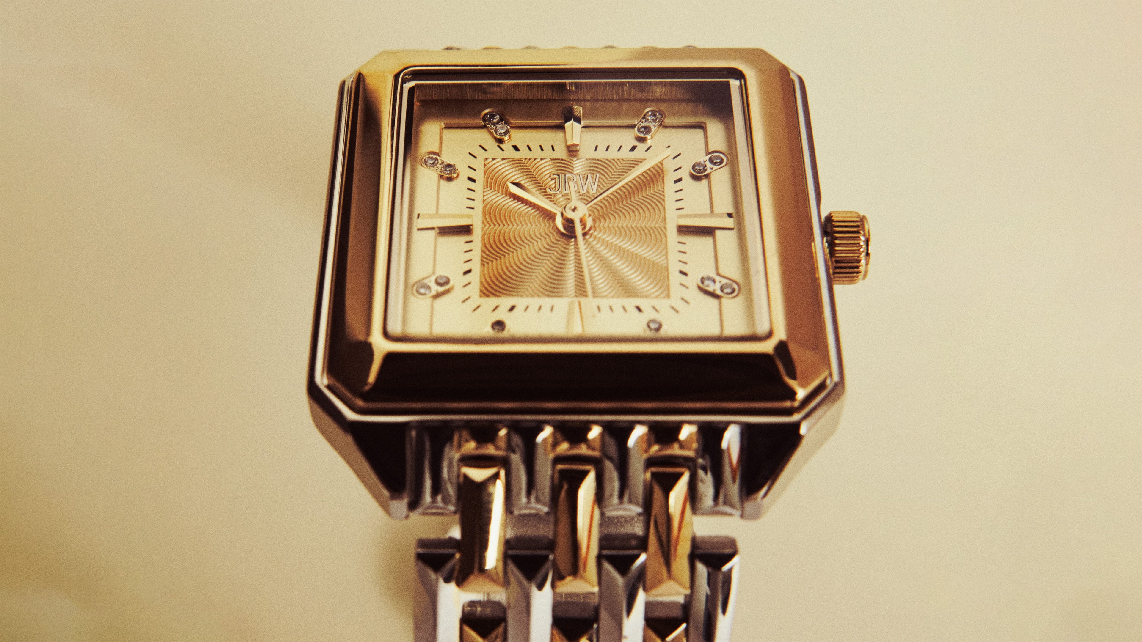 A close-up of a square gold wristwatch with a textured face, metallic hour markers, and a linked metal band against a neutral background.