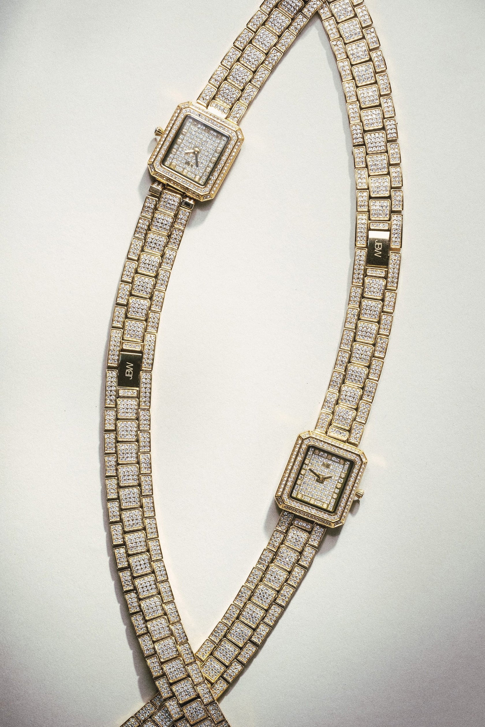 Two JBW Watches Arc Double (Sample Sale) | J6390C-GA (Grade A) gold wristwatches with square faces and crystal-studded bezels are arranged in a crisscross on a light background, highlighting their elegant design.