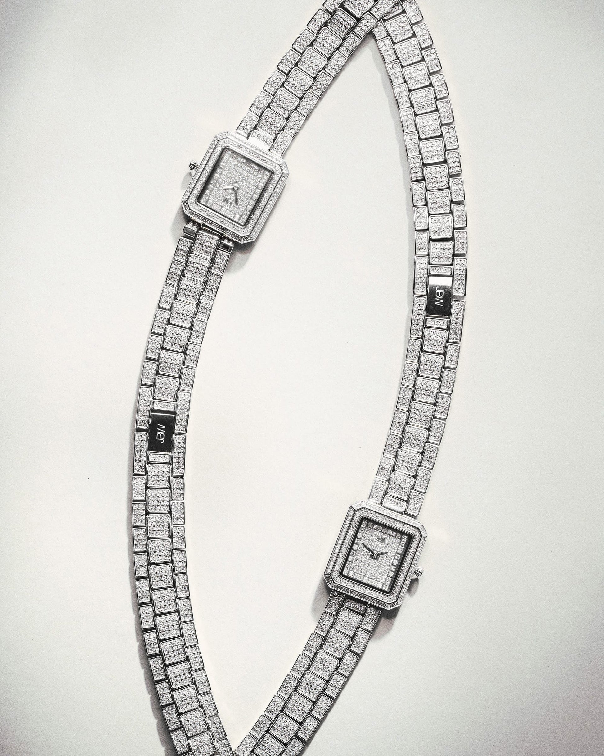 Two silver, square-faced JBW Watches Arc Double | J6390D wristwatches with sparkling metal bands are arranged in an S-curve on a light background. Each stylish timepiece displays a different time and features matching glittering designs.
