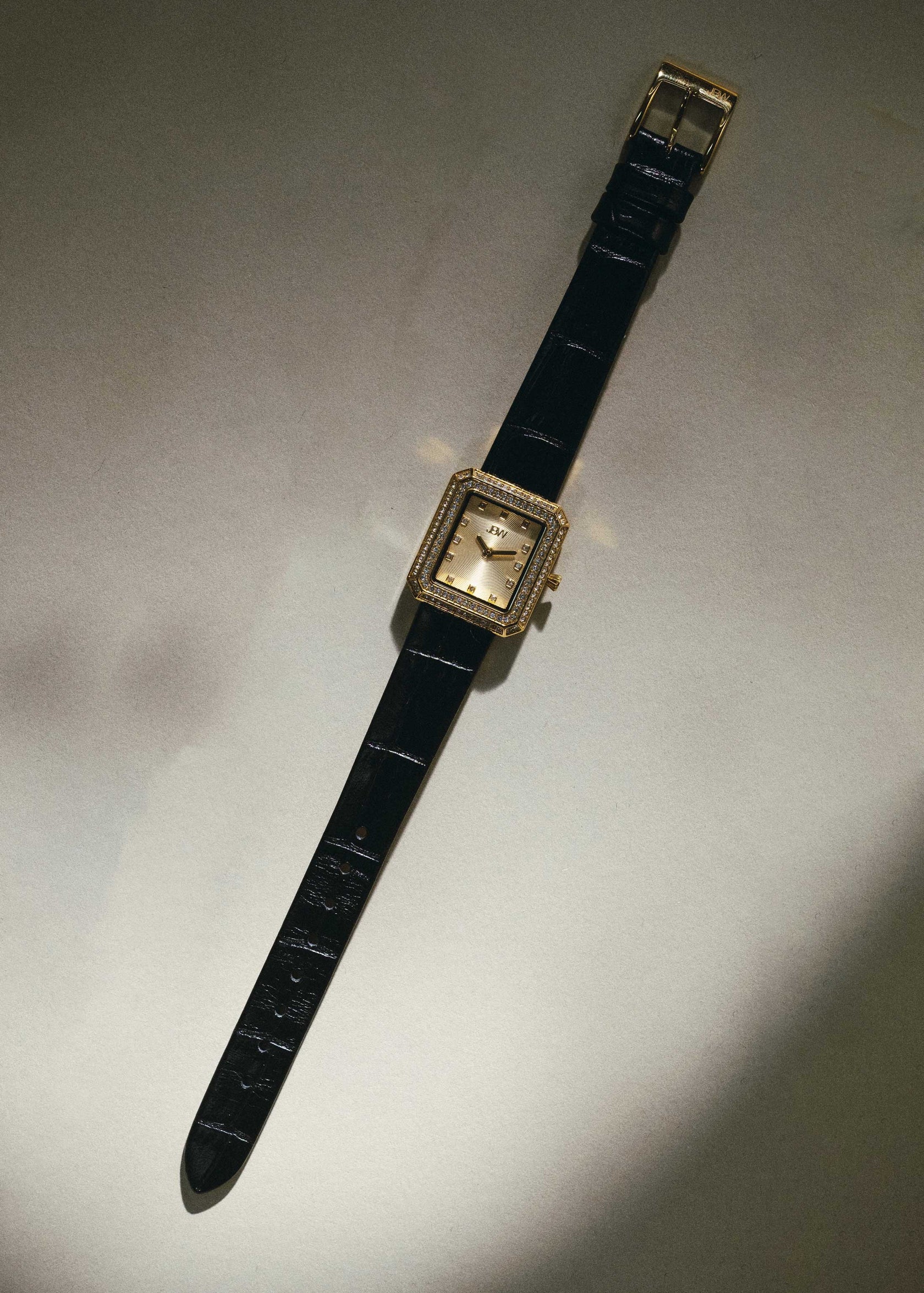 The JBW Watches Arc Leather | J6390LA is a gold rectangular unisex watch with a diamond-encrusted bezel and black leather band, displayed flat on a neutral surface under soft lighting.