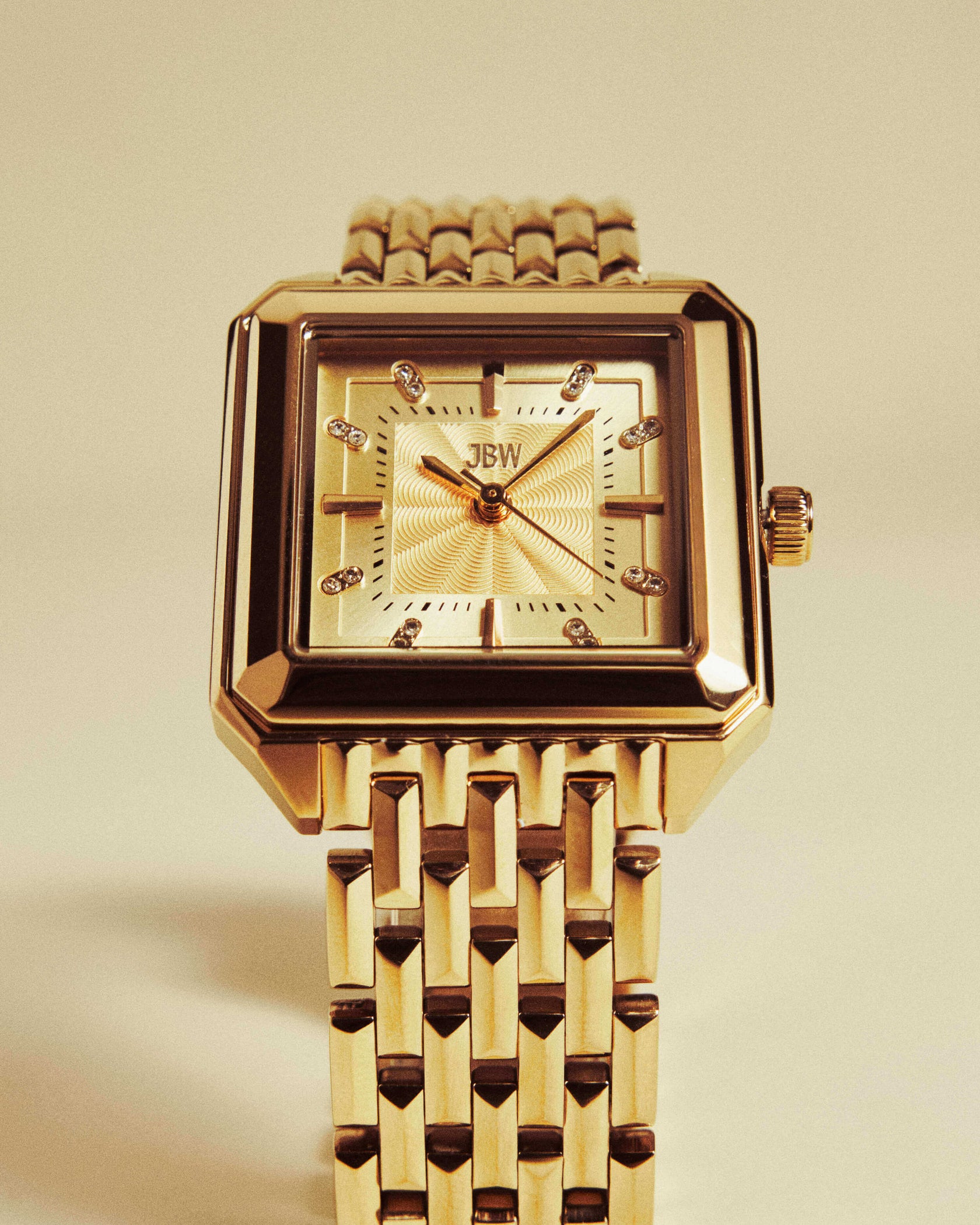 The JBW Watches Astra | J6410B is a gold square silhouette watch with a metal link band, diamond hour markers, Swiss quartz movement, and a guilloché-textured dial, shown on a light beige background.