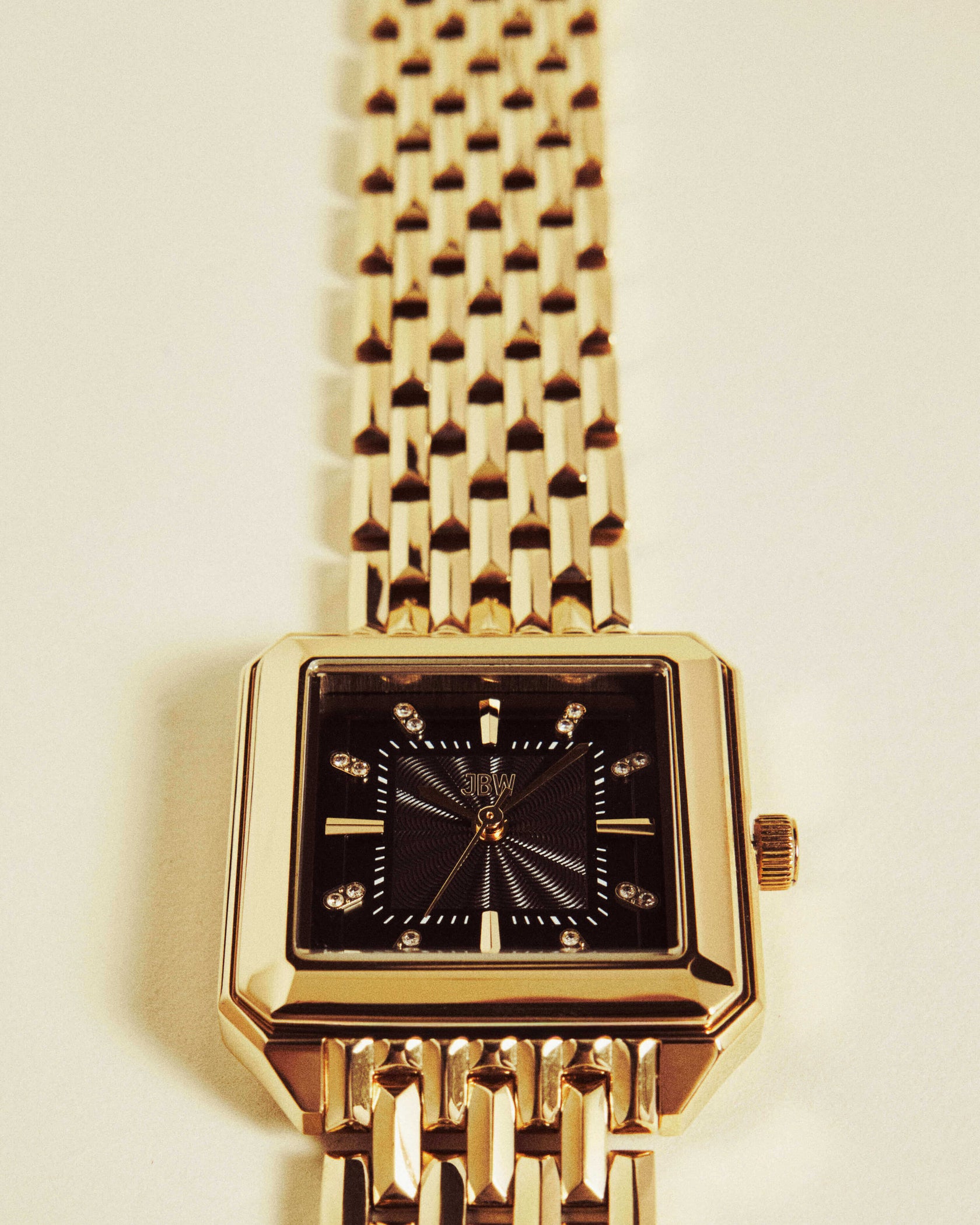 The JBW Watches Astra | J6410D, a gold wristwatch with a guilloché square black dial, gold hour markers, diamond-like accents at 3, 6, 9, and 12 o’clock, and a linked gold band is laid flat on a light surface.