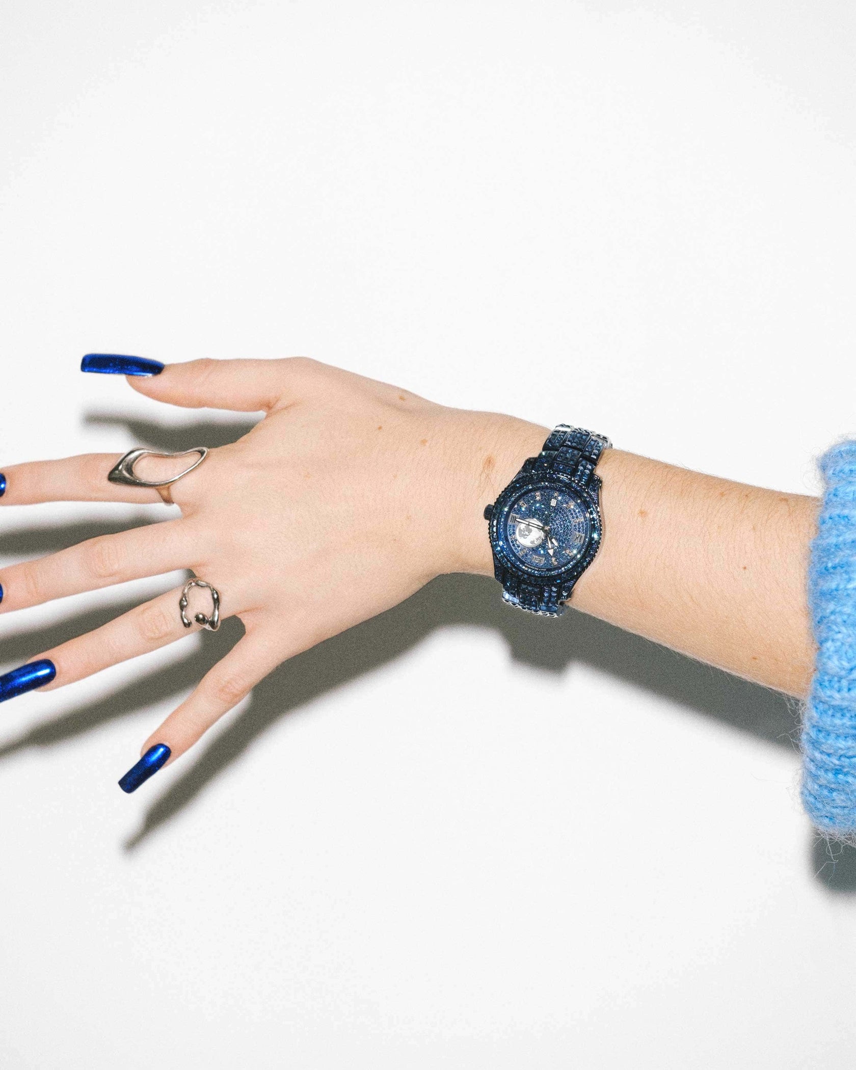 A hand with blue painted nails and silver rings wears a JBW Watches RESERVE Bleu 39 | R6393A-GA (Grade A) watch, shown against a white background with a blue sweater sleeve visible.