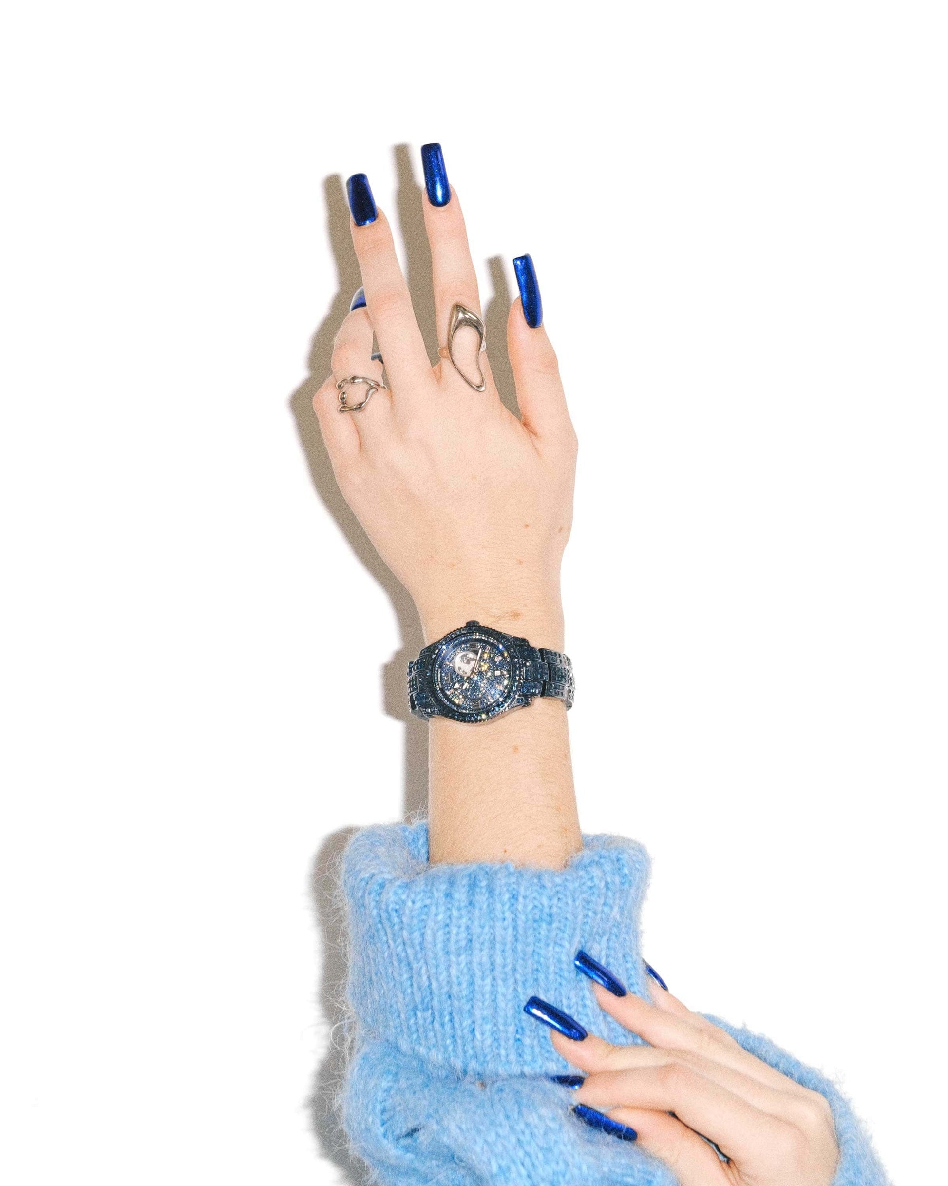 A hand adorned with the JBW Watches RESERVE Bleu 39 | R6393A-GA sparkly dark watch and silver rings, featuring long blue nails, is raised against a white background. The wearer is dressed in a fuzzy light blue sweater.