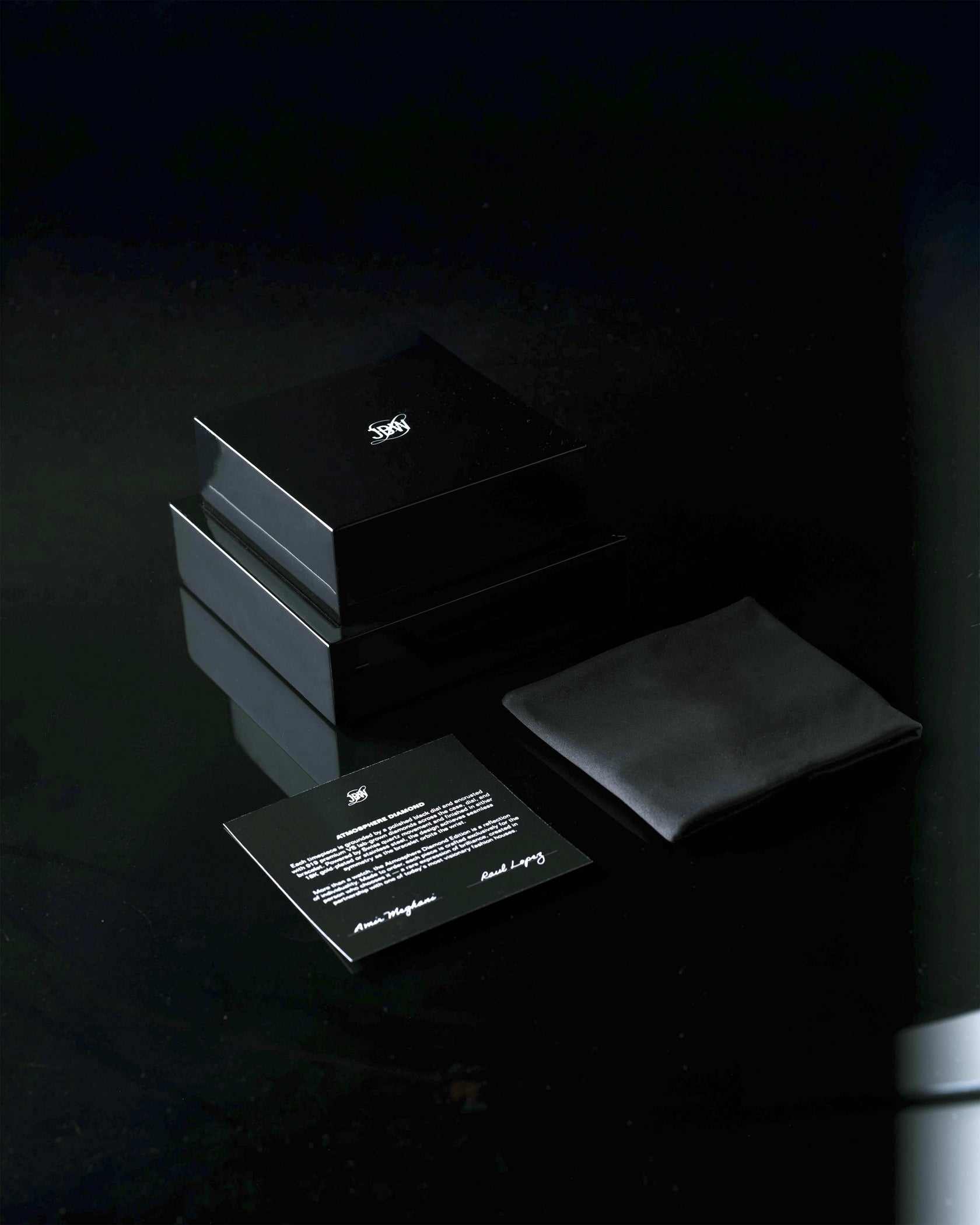 Two glossy black boxes, a black cloth pouch, and an instruction card with a white emblem from JBW Watches are arranged on a reflective surface, hinting at the luxury JBW x Luar – Atmosphere Diamond | J6407A timepiece within.