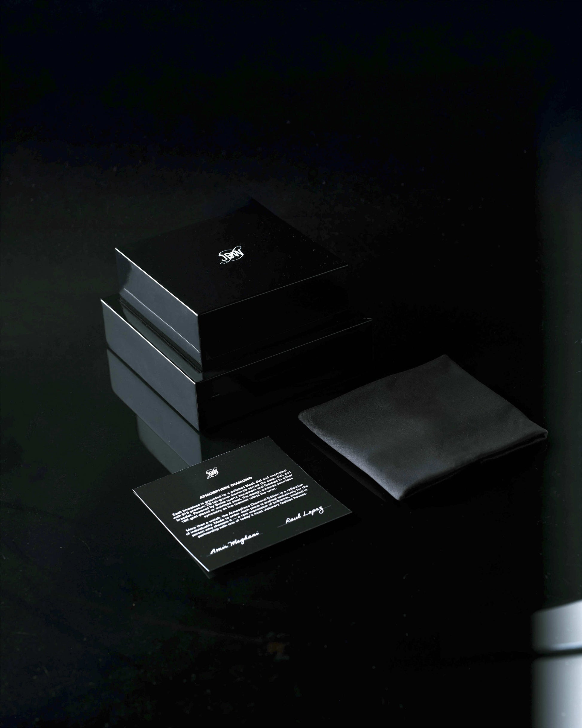 Two glossy black boxes, a black cloth pouch, and an instruction card with a white emblem from JBW Watches are arranged on a reflective surface, hinting at the luxury JBW x Luar – Atmosphere Diamond | J6407A timepiece within.