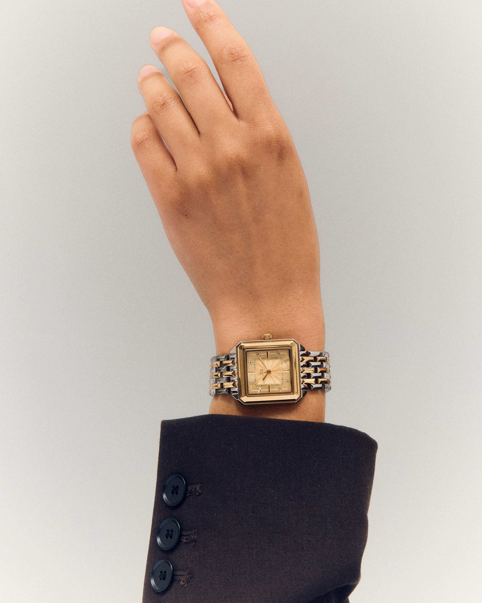 A person in a dark brown suit raises their hand, displaying the JBW Watches Astra | J6410C—a square watch with Swiss quartz movement and a gold stainless steel bracelet—against a light, neutral background.