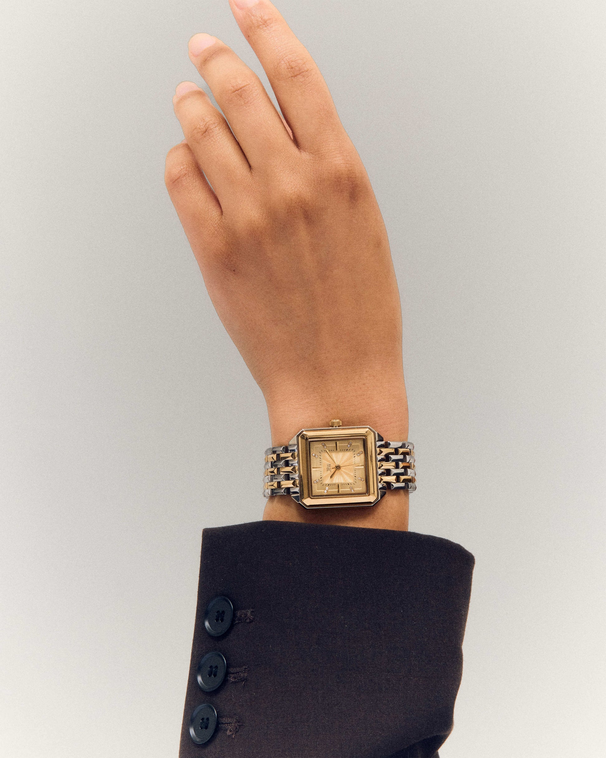 A person in a dark brown suit raises their hand, displaying the JBW Watches Astra | J6410C—a square watch with Swiss quartz movement and a gold stainless steel bracelet—against a light, neutral background.
